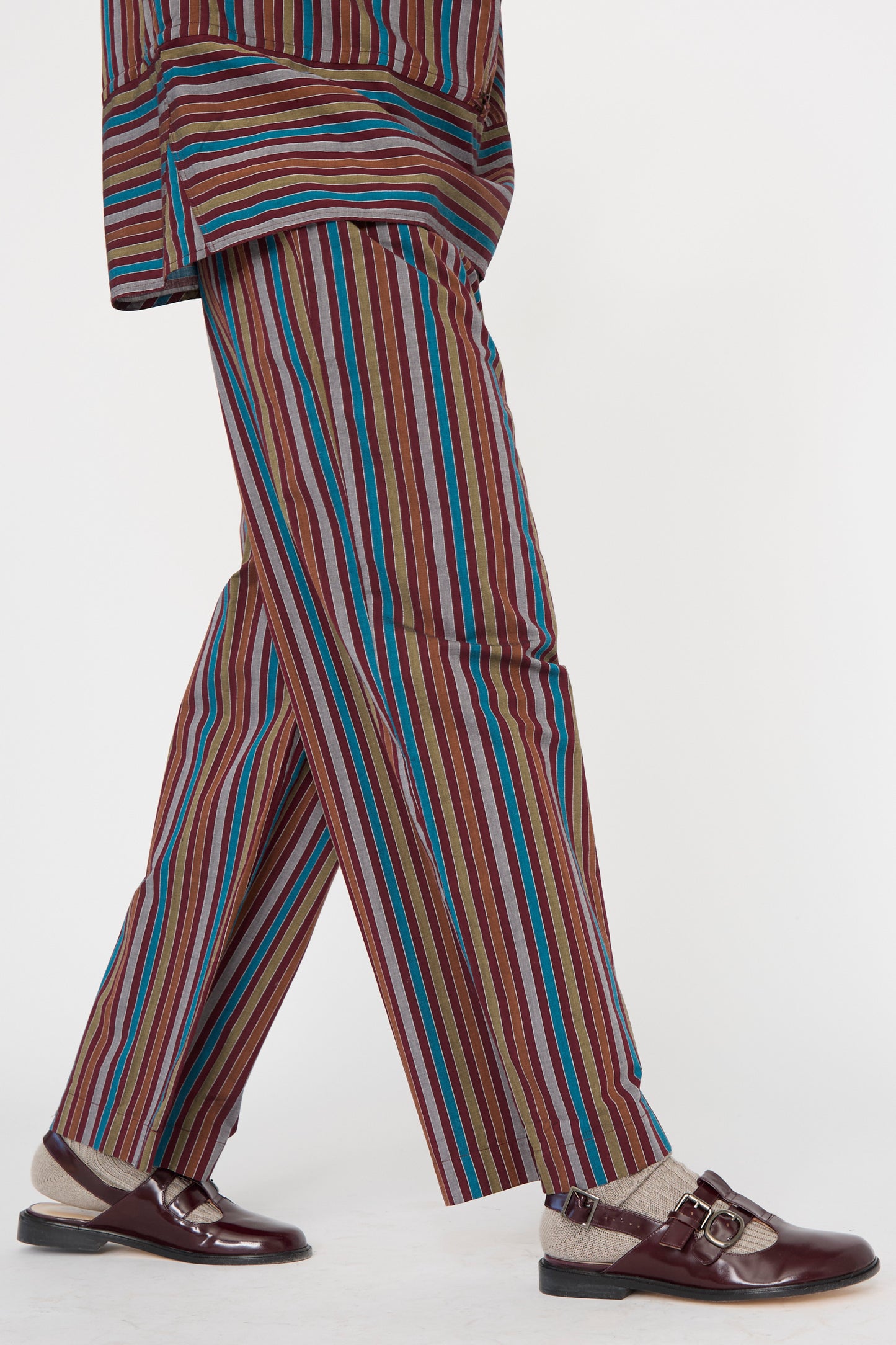 A person wearing Baserange Cotton Poplin Ole Pants in Maroon Multi Stripe with dark brown shoes and beige socks against a plain backdrop. 