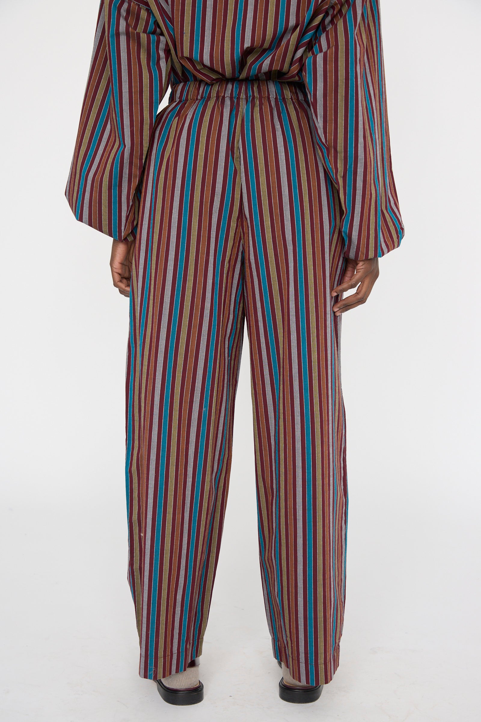 A person in Baserange's Cotton Poplin Ole Pants in Maroon Multi Stripe presents a back view against a plain backdrop. The relaxed-fit, wide-leg striped organic cotton pants flaunt vertical red, blue, and brown stripes.