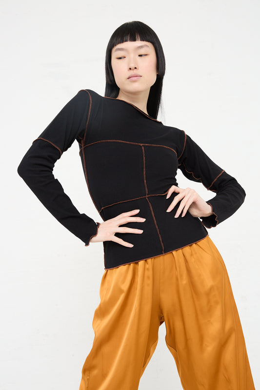 A woman standing with hands on hips wearing a Baserange Organic Cotton Rib Cinder Long Sleeve in Black with orange stitching, and mustard-colored trousers against a white background.