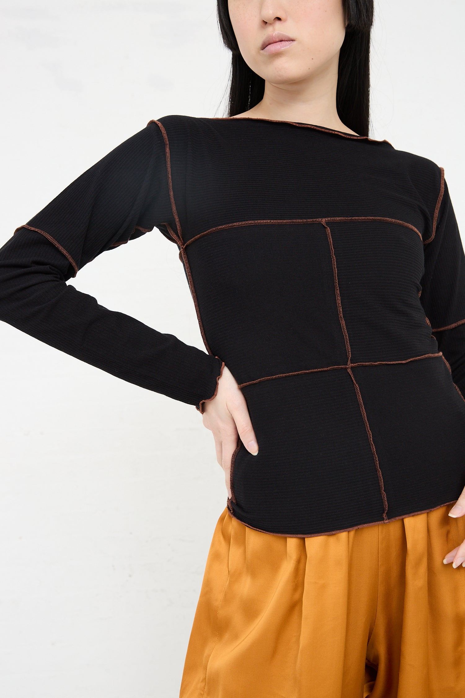 Woman wearing a Baserange Organic Cotton Rib Cinder Long Sleeve in Black with brown stitching, posing with hand on hip against a white background.