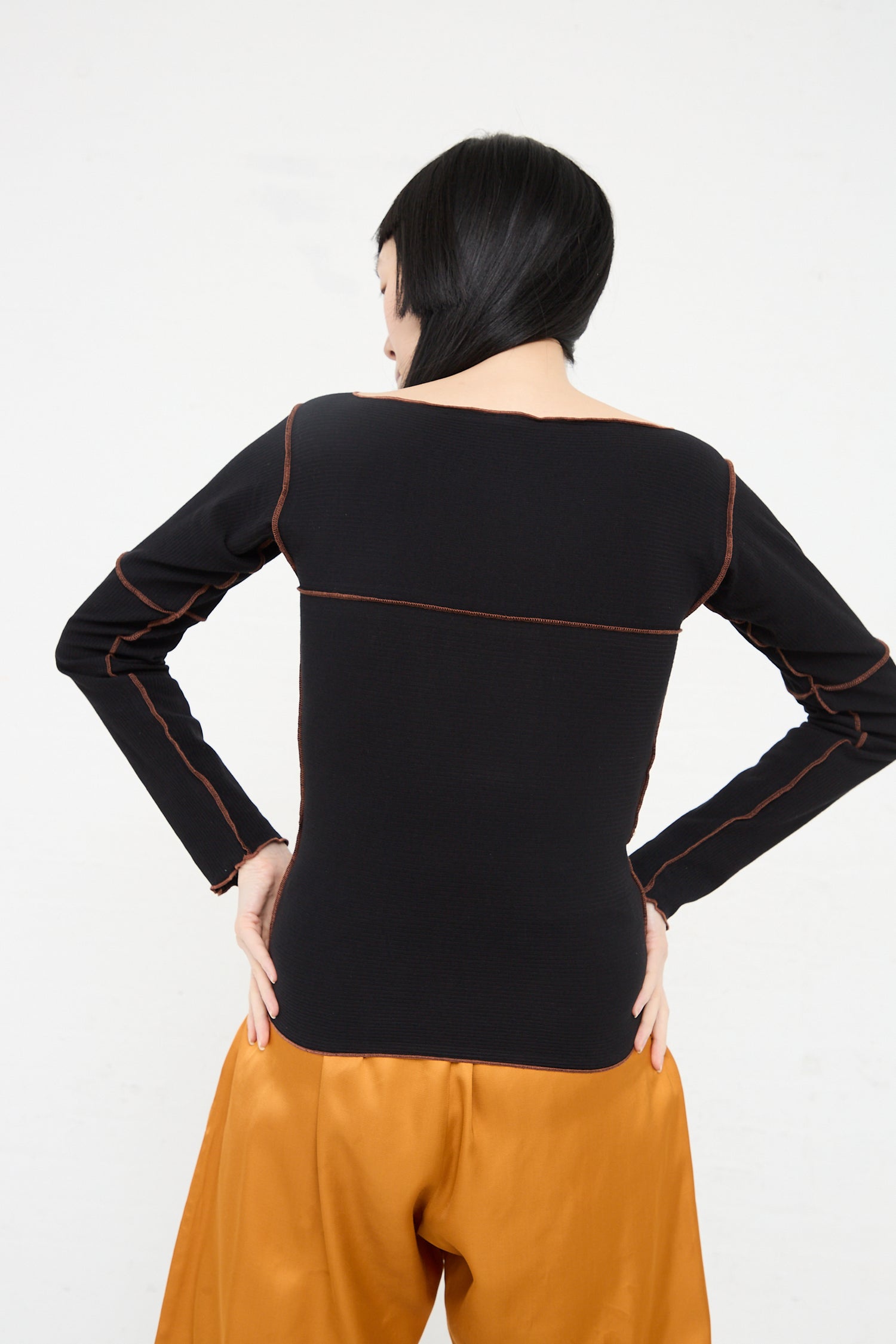 Woman with bob haircut, standing with back to camera, wearing a Baserange Organic Cotton Rib Cinder Long Sleeve in Black with orange stitching and orange pants.