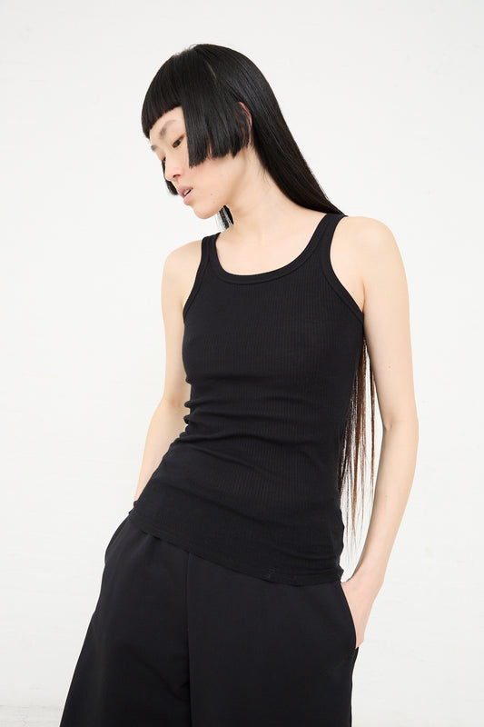 A woman with long black hair and bangs, wearing a Baserange Organic Cotton Rib Drive Tank in Black and trousers, posing against a white background.