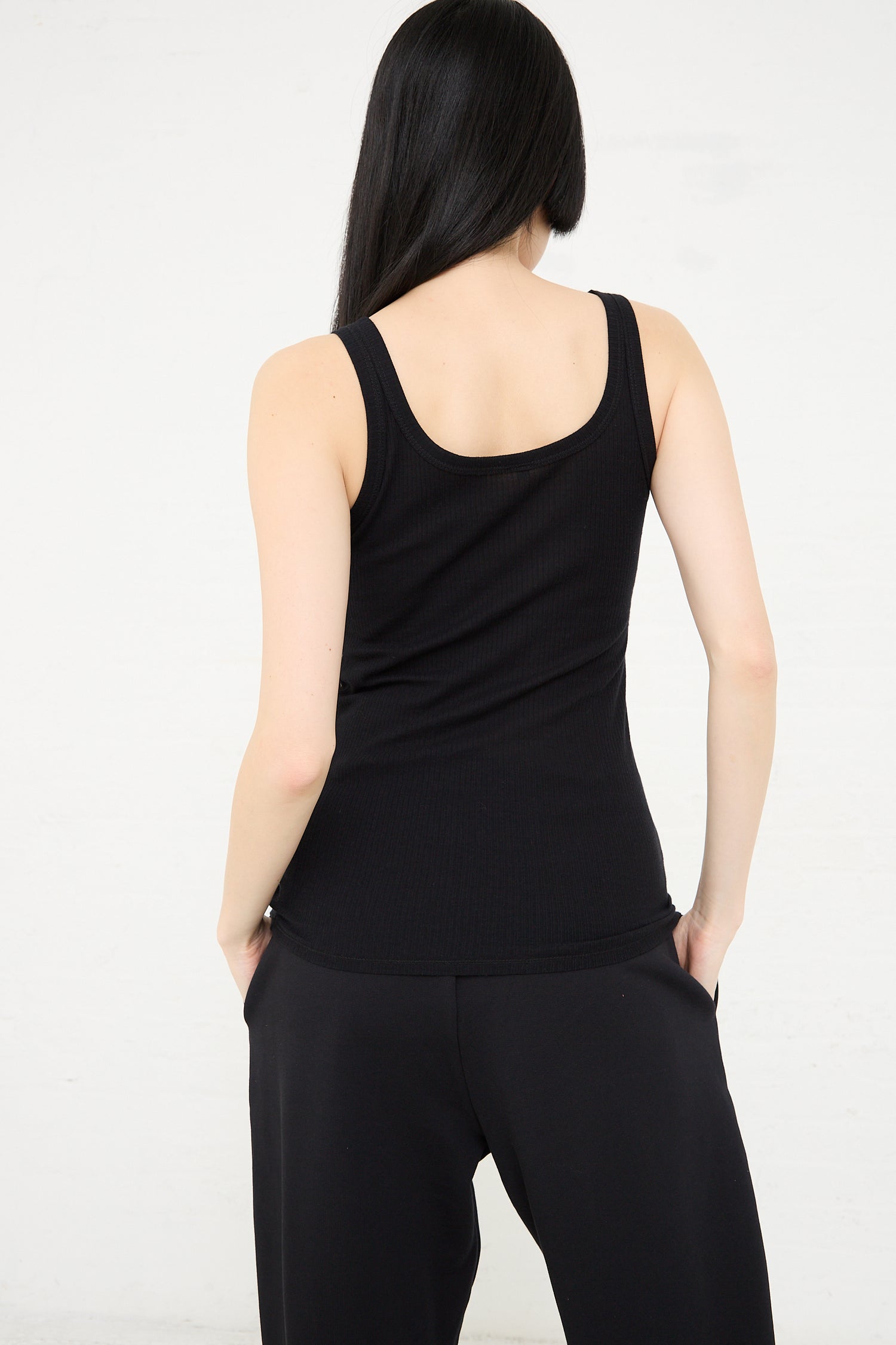 Woman from behind, wearing a Baserange Organic Cotton Rib Drive Tank in Black and black pants, standing against a white background.