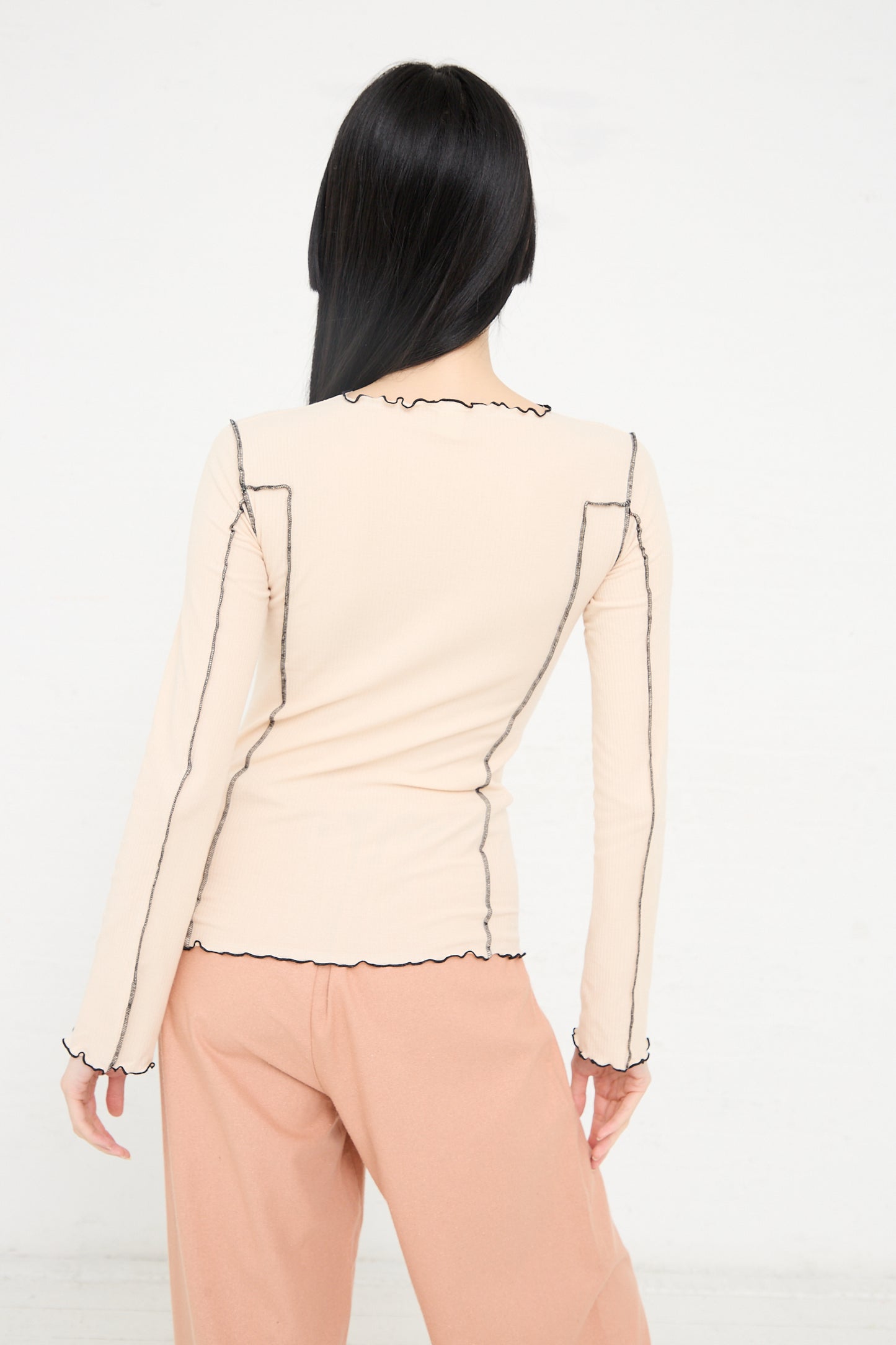 Woman with long black hair, seen from behind, wearing a beige boatneck top with black stitched details and peach pants against a white background - Organic Cotton Rib Omato Long Sleeve in Kano Pink by Baserange.