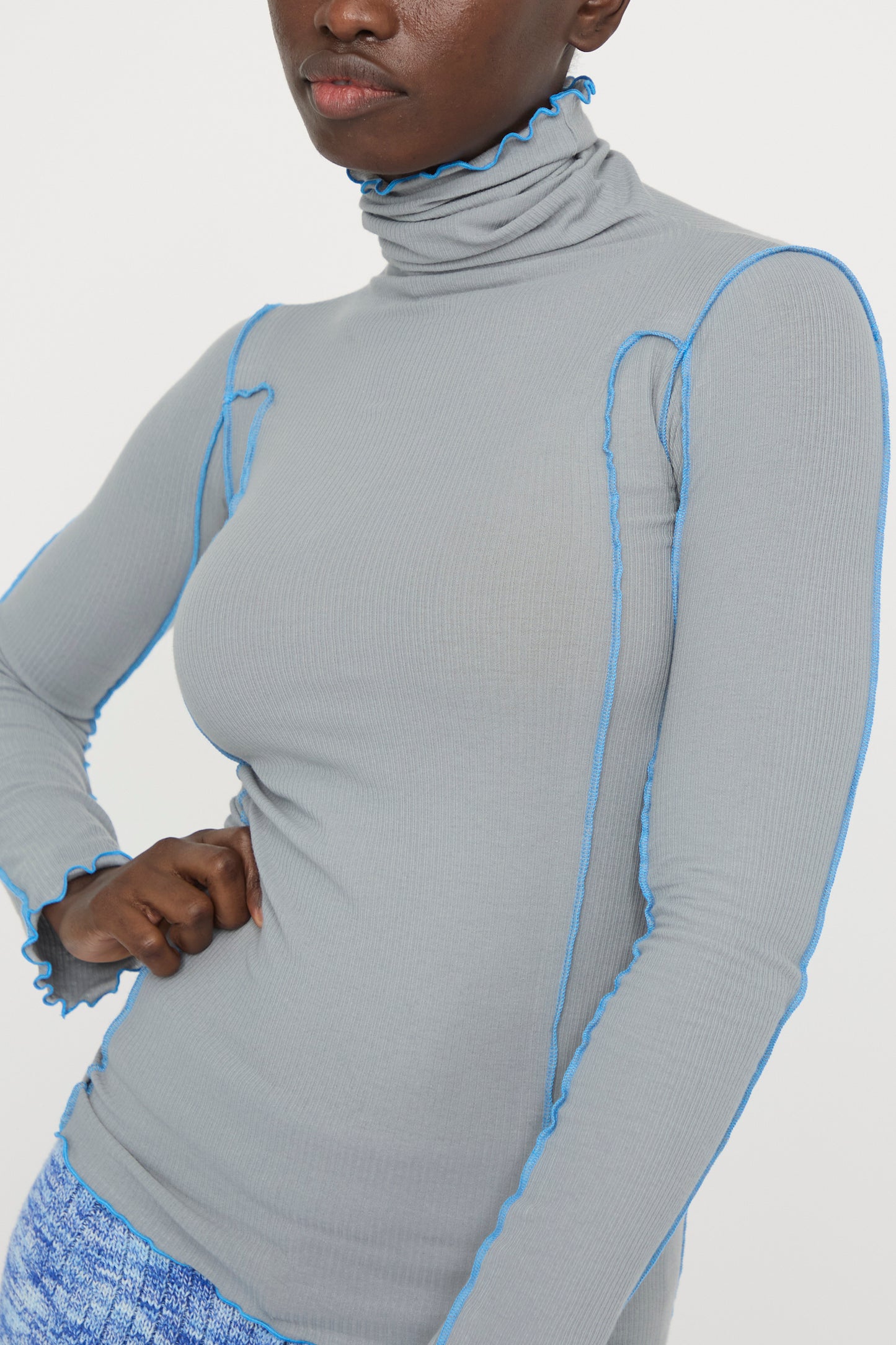 A person stands with a hand on their hip wearing the Bow Blue Cotton Rib Omato Turtleneck by Baserange against a white background. 