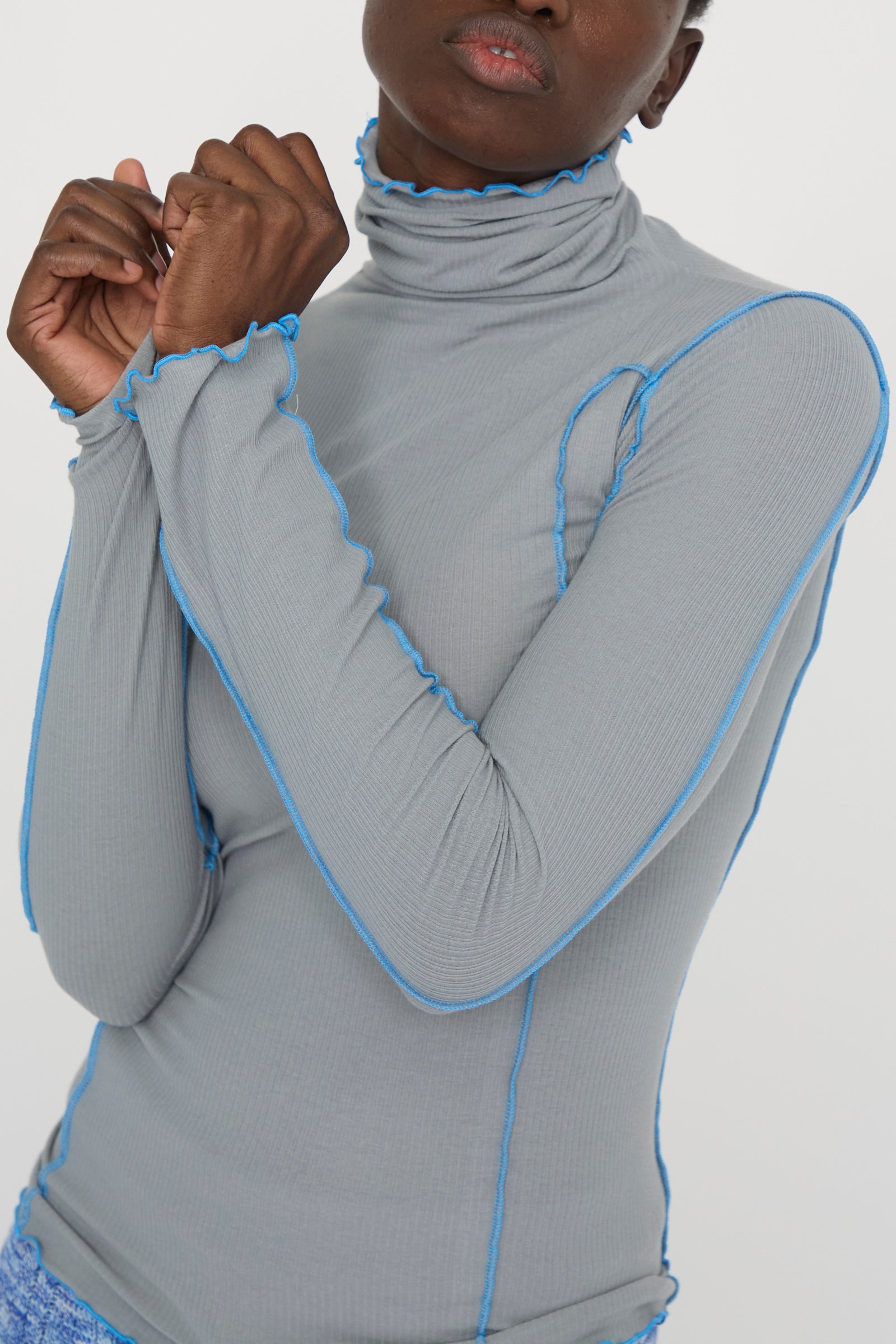 A model wearing the Baserange Cotton Rib Omato Turtleneck in Bow Blue, featuring gray fabric with blue stitching and crafted from organic cotton, holds their hands together.