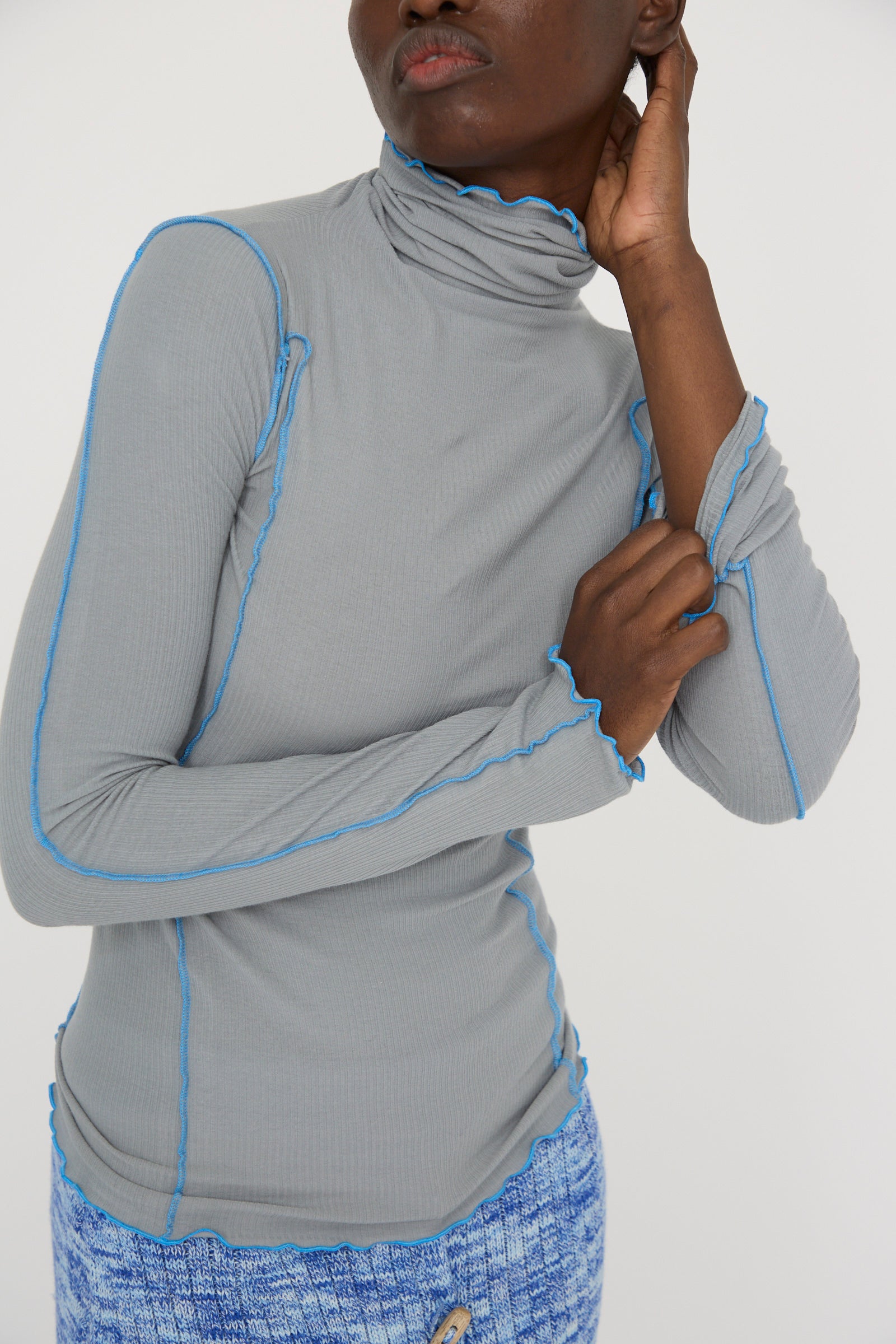 A model wearing Baserange's Cotton Rib Omato Turtleneck in Bow Blue and a textured organic cotton skirt stands against a plain background, touching their head with one hand and holding the sleeve with the other.