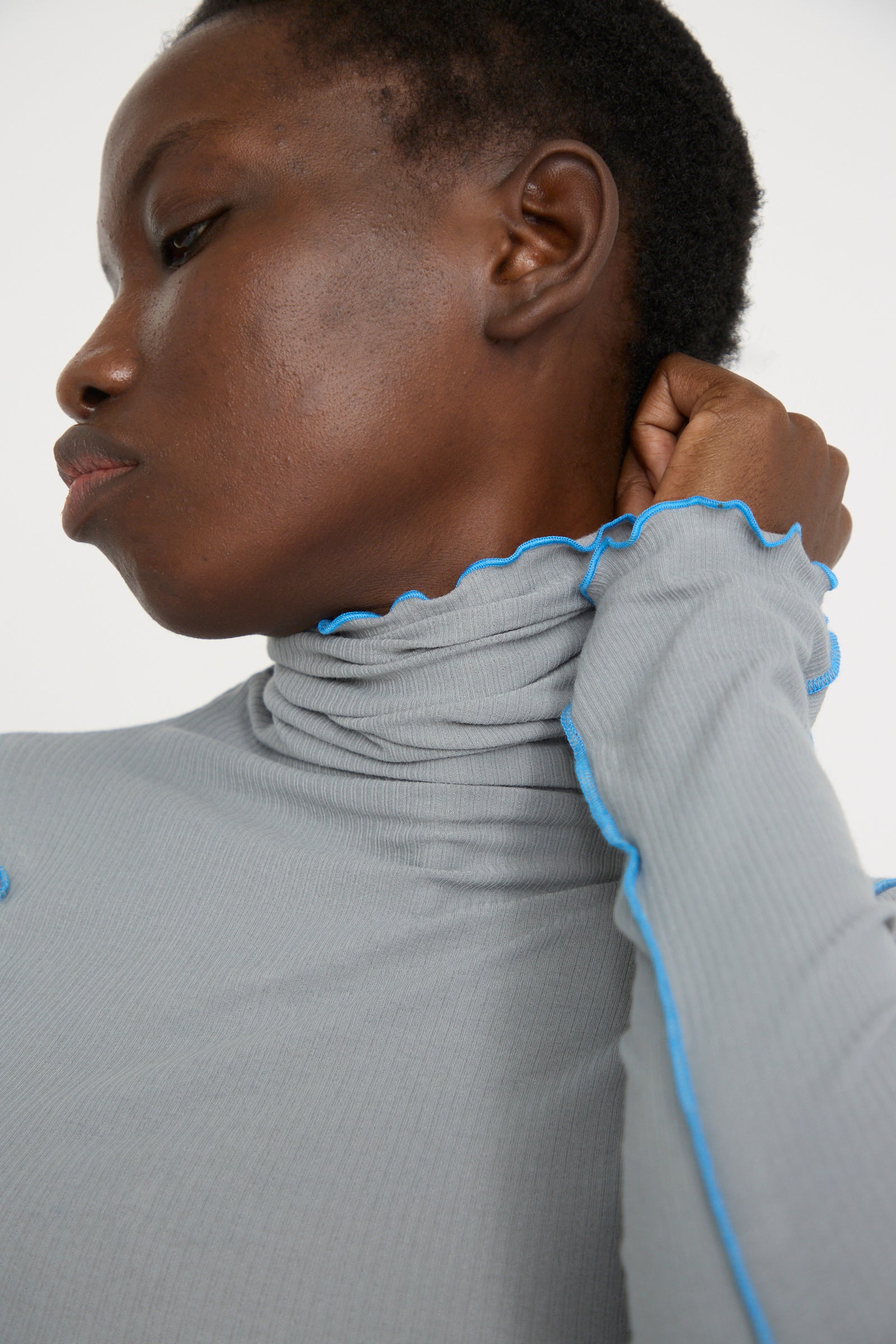 The individual is adjusting the collar of a Baserange Cotton Rib Omato Turtleneck in Bow Blue, crafted from organic cotton, and gazing sideways.