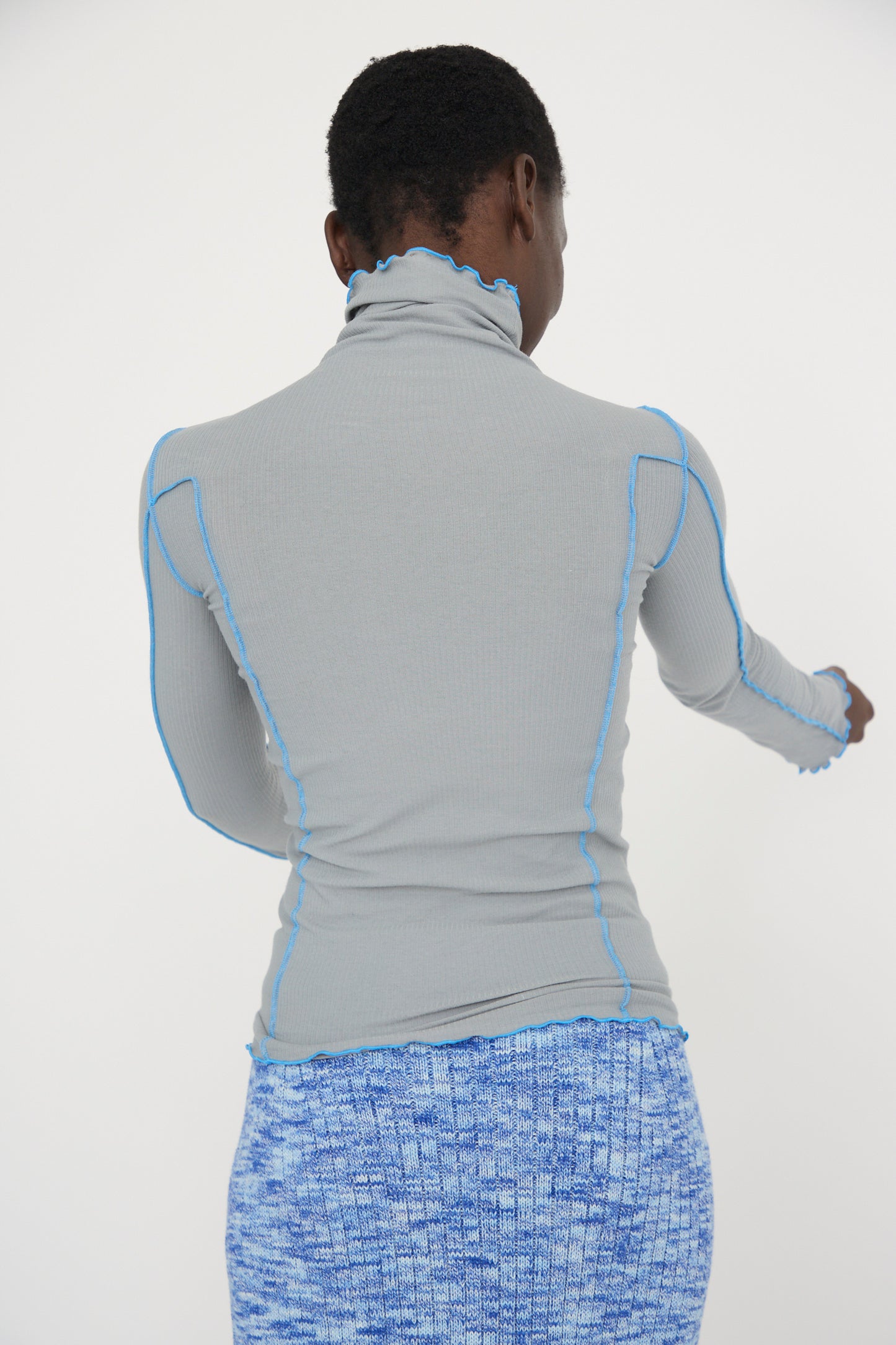 A person wearing the Baserange Cotton Rib Omato Turtleneck in Bow Blue and a blue patterned skirt is standing with their back facing the camera against a white backdrop. 