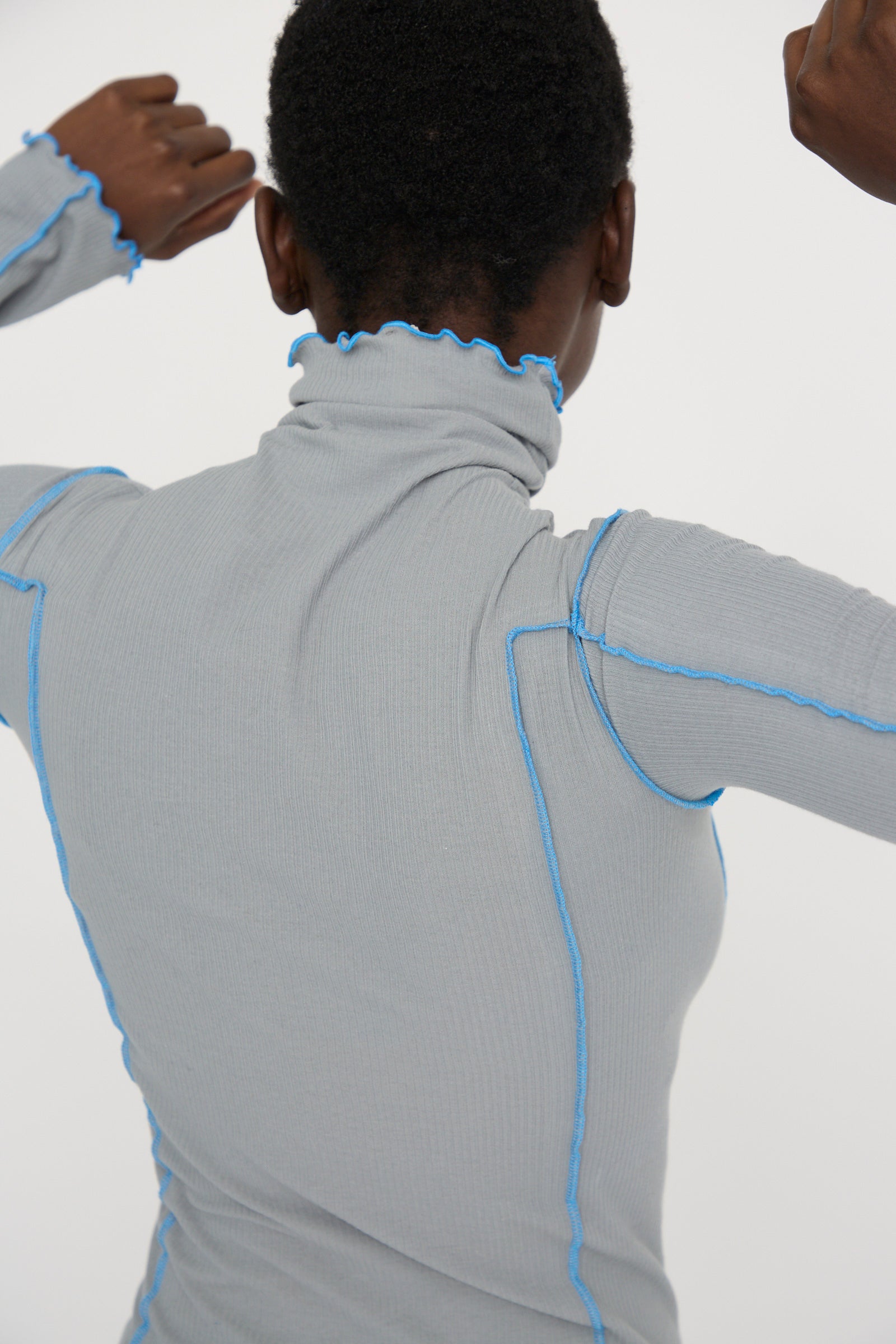 A person wearing the Baserange Cotton Rib Omato Turtleneck in Bow Blue, featuring gray organic cotton with blue seams and ruffled edges, stands with their back to the camera.