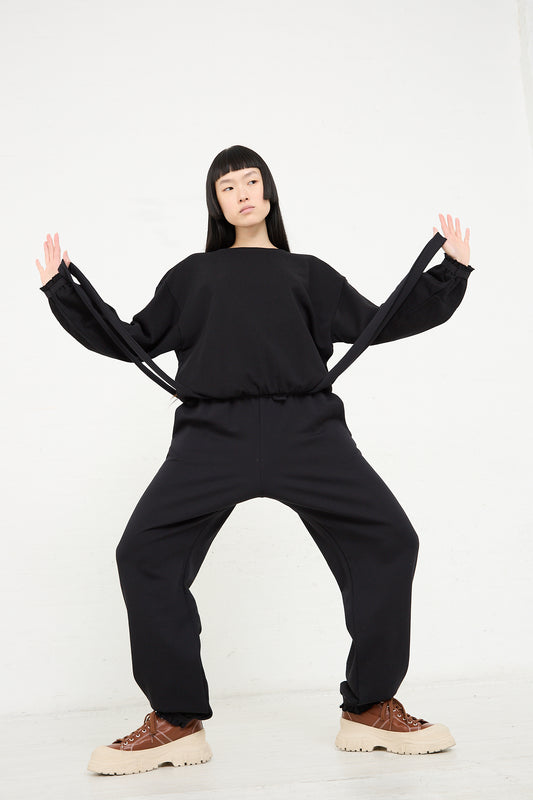 A woman in a Baserange Organic Cotton Route Sweatshirt in Black and beige shoes stands against a white background, posing with arms extended and hands facing outward.