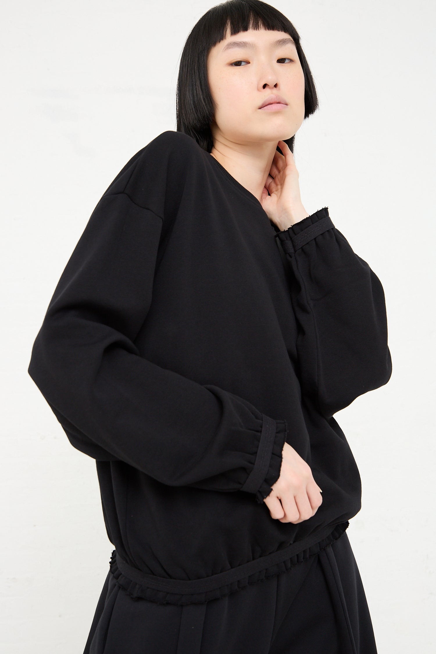 Woman in a Baserange Organic Cotton Route Sweatshirt in Black and pants posing with her hand on her neck, against a white background.