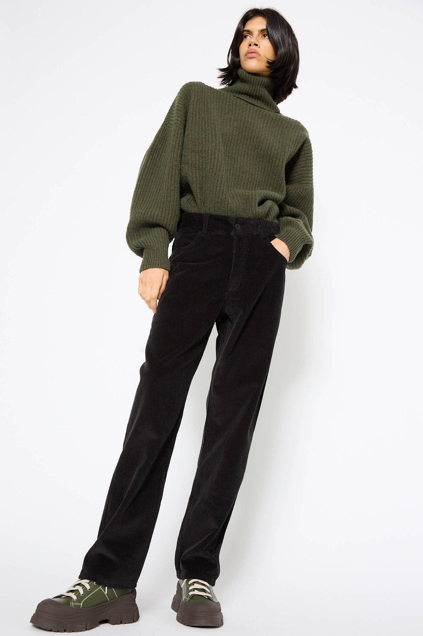 A person in a green turtleneck sweater and Baserange's Cotton Tajo Pant in Black stands against a white background, hands in pockets, wearing green and brown sneakers.