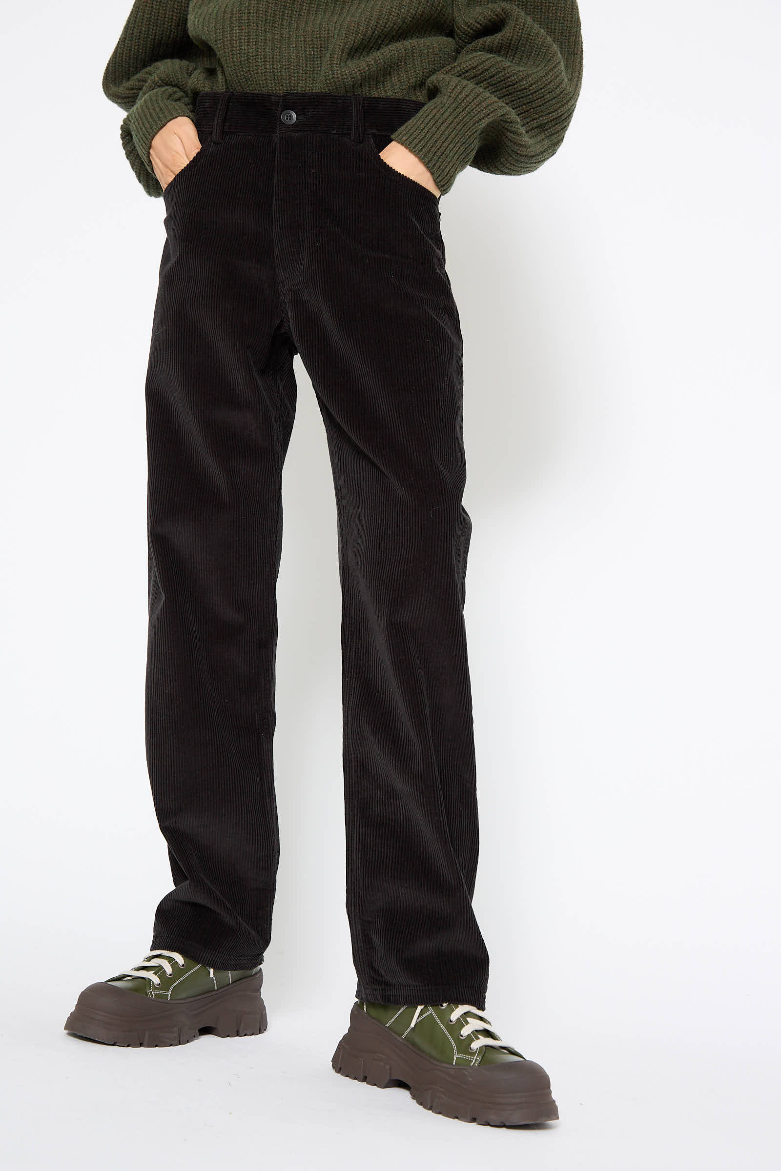 Person wearing Baserange Cotton Tajo Pant in Black, a green sweater, and green lace-up shoes stands against a white background.