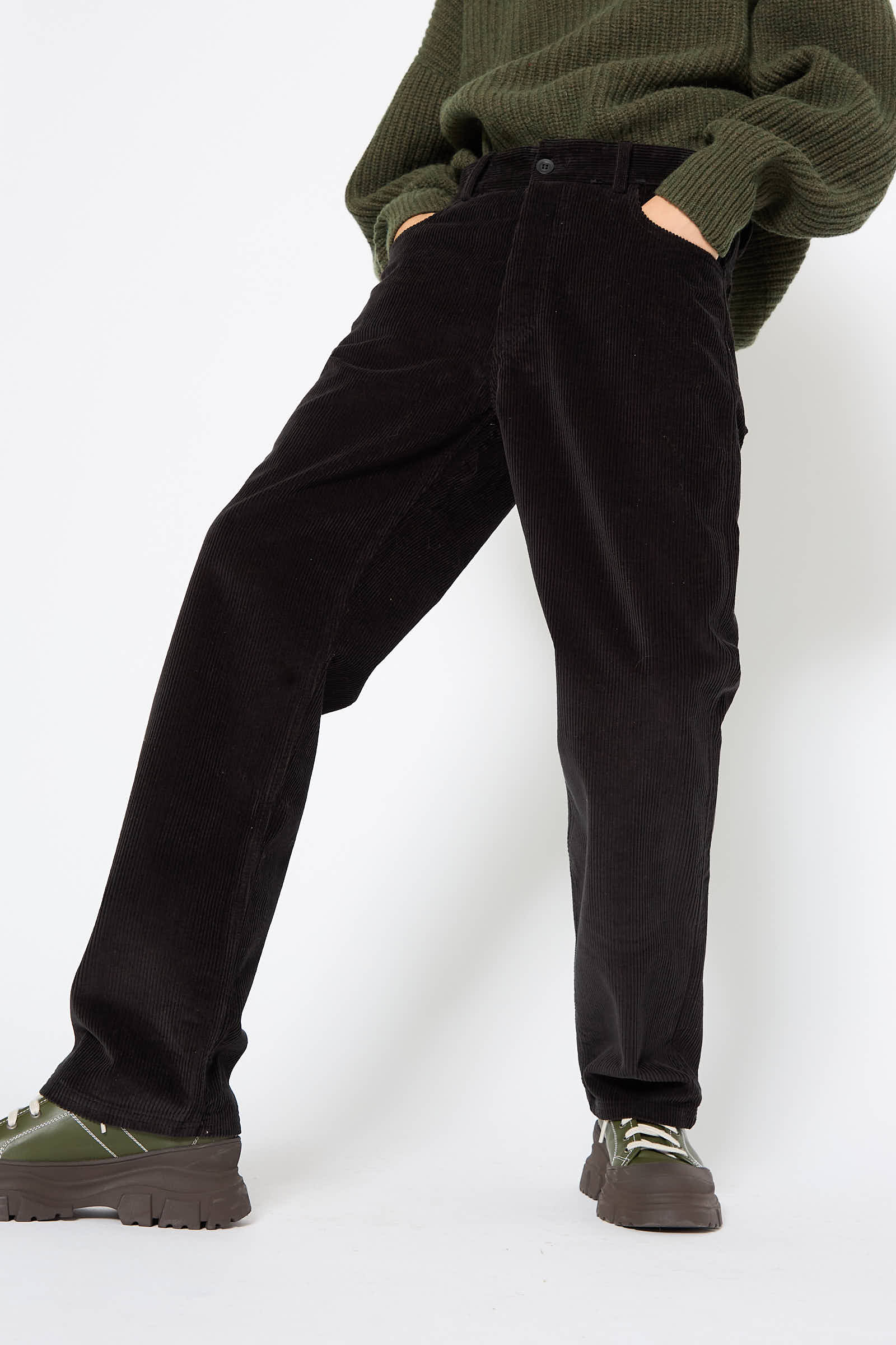 Person wearing Baserange's Cotton Tajo Pant in Black, green sweater, and green sneakers standing with hands in pockets against a plain background.