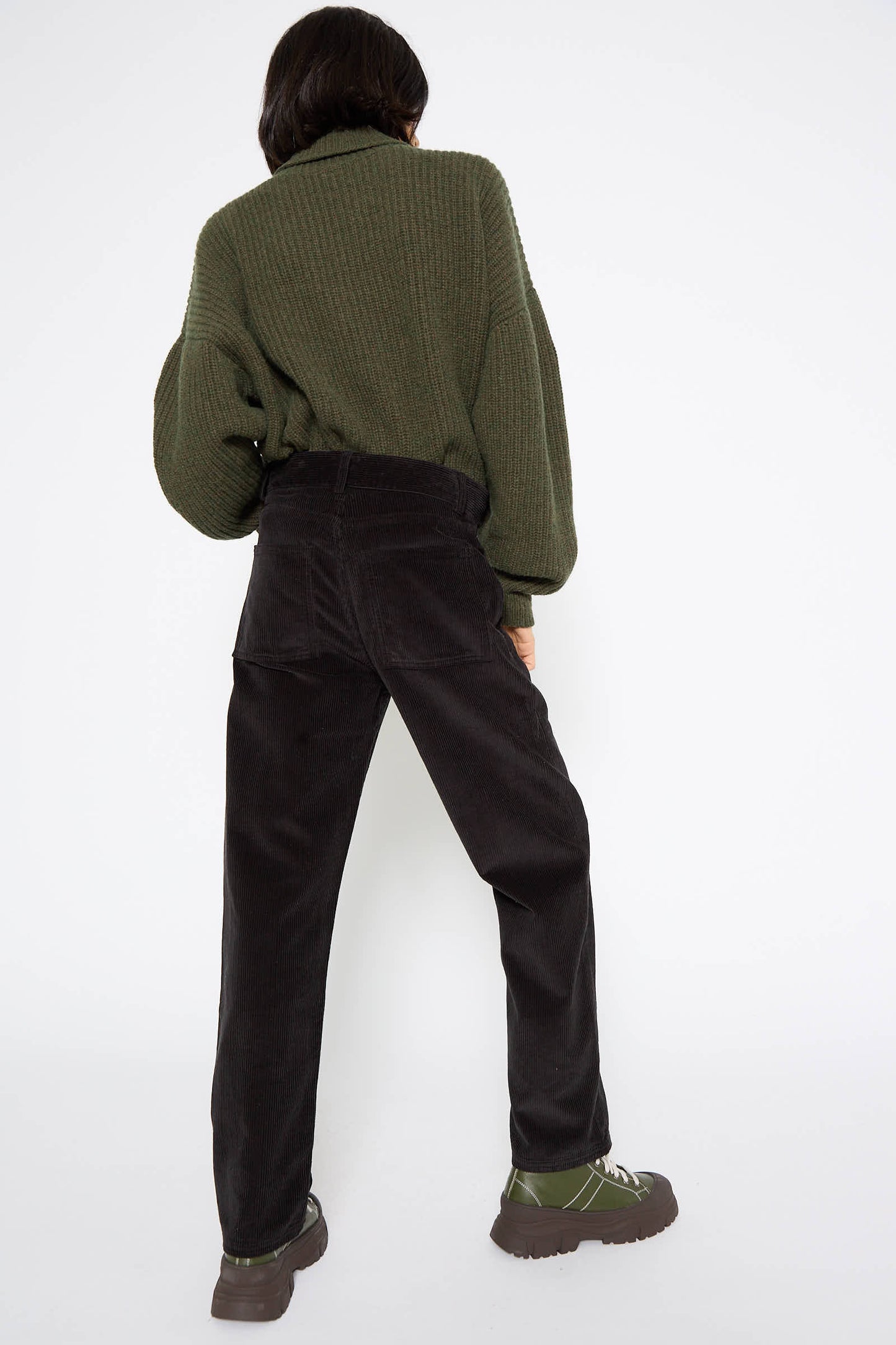 A person stands facing away, wearing a green sweater, Cotton Tajo Pant in Black by Baserange in a relaxed fit, and green shoes against a plain white background.