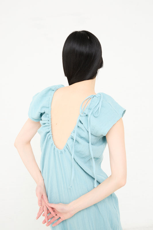 Woman with her back turned to the camera, wearing a Crinkle Linen Cotton Max Dress in Wuxi Blue by Baserange, with an open back and a bow detail at the top.