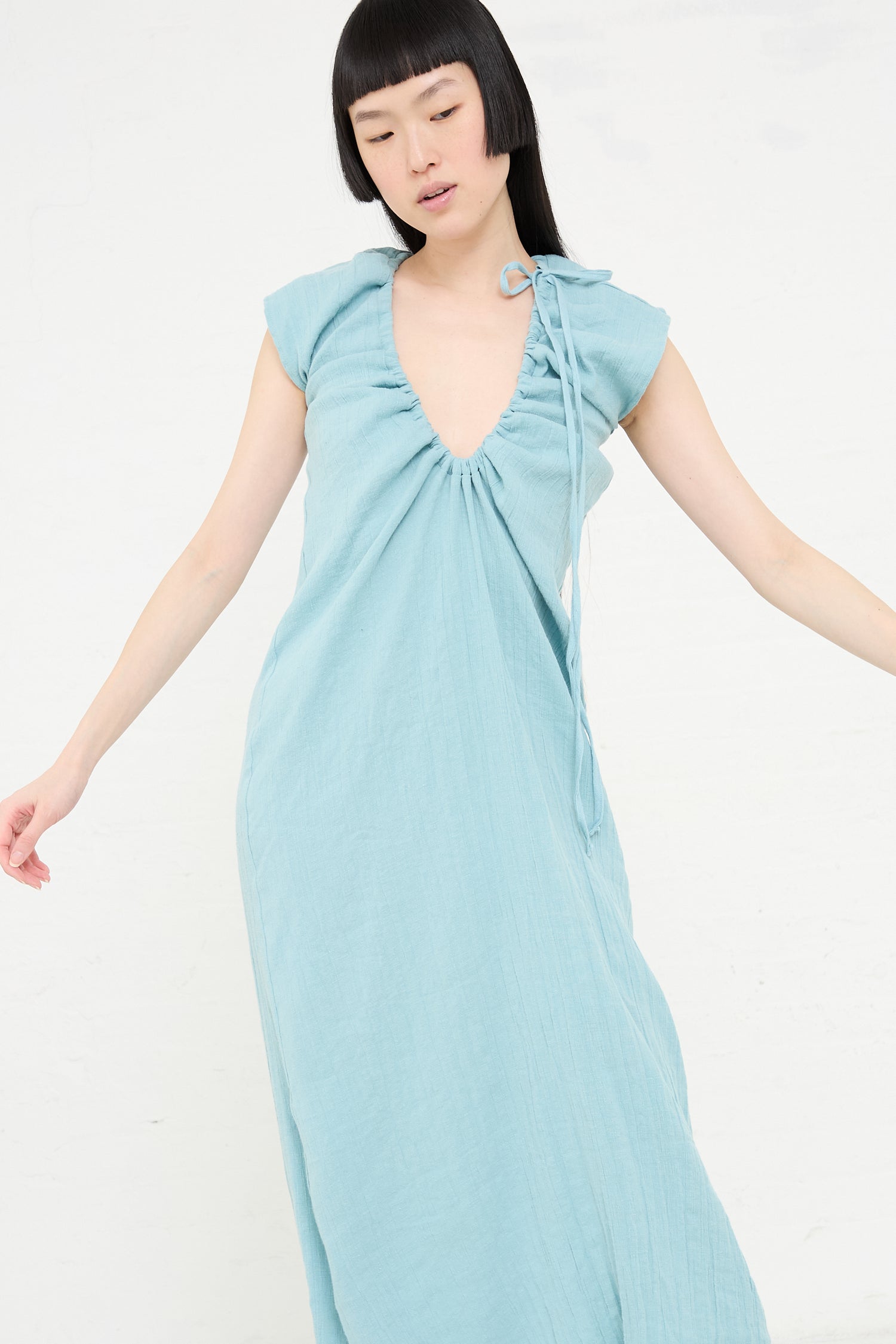 Woman posing in a Crinkle Linen Cotton Max Dress in Wuxi Blue by Baserange, with a tied shoulder detail.
