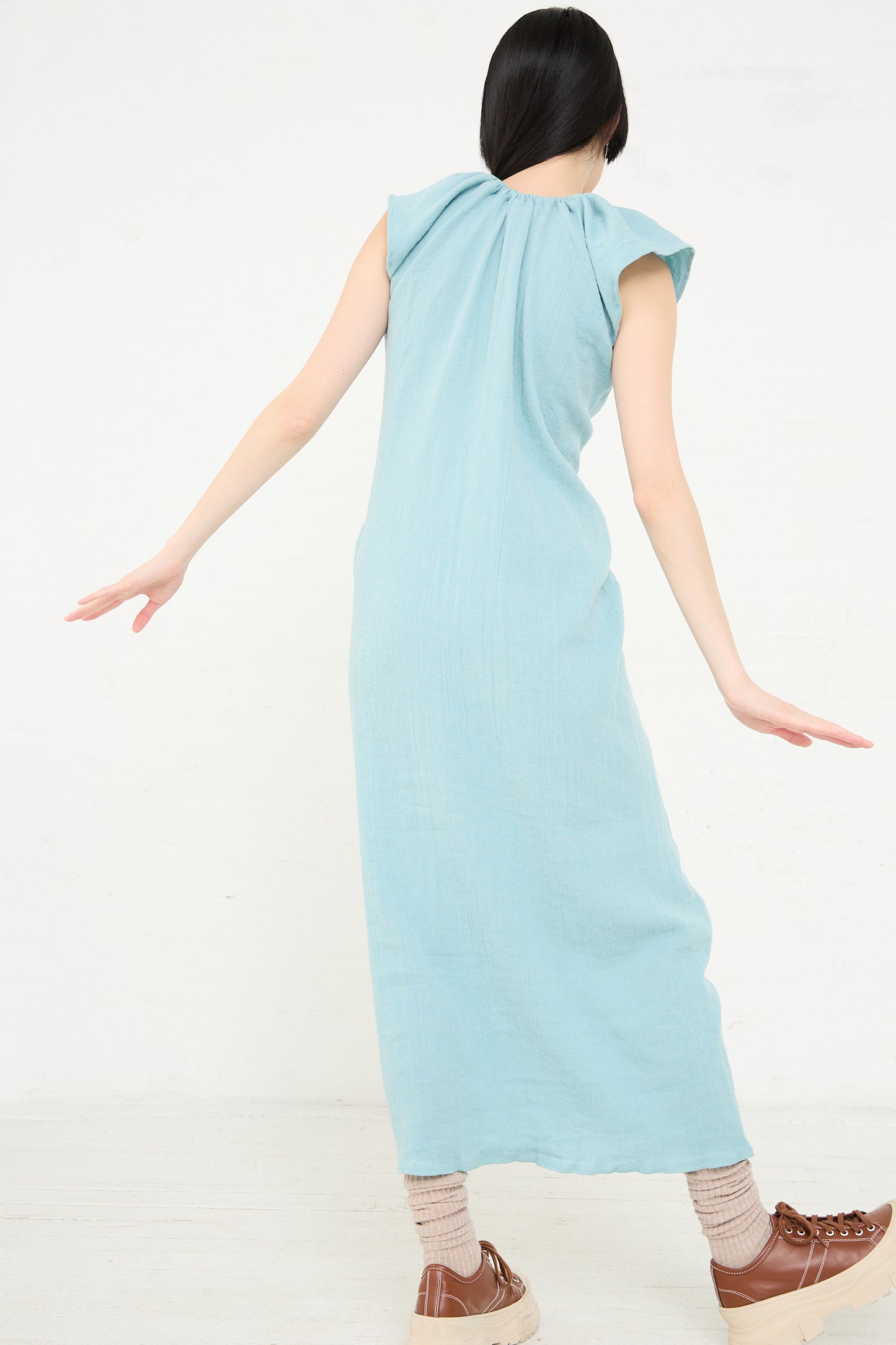 A woman seen from behind, wearing a Crinkle Linen Cotton Max Dress in Wuxi Blue by Baserange and brown shoes, is standing with her arms spread out to the sides against a plain background.