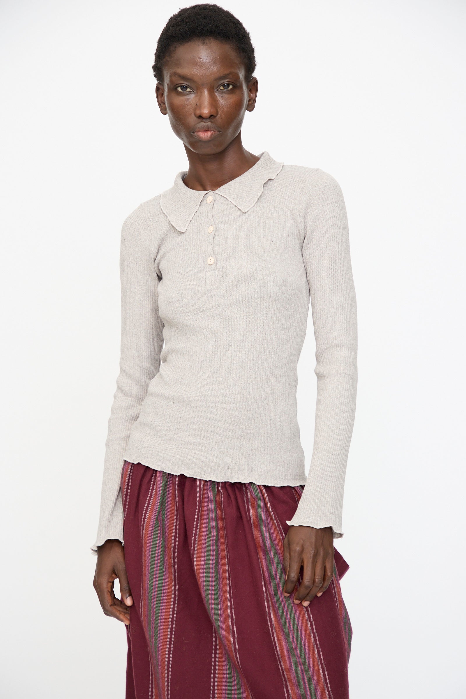 A person stands against a plain background wearing the Baserange Dual Long Sleeve Tee in Grey Melange, paired with a maroon skirt featuring vertical multicolored stripes.