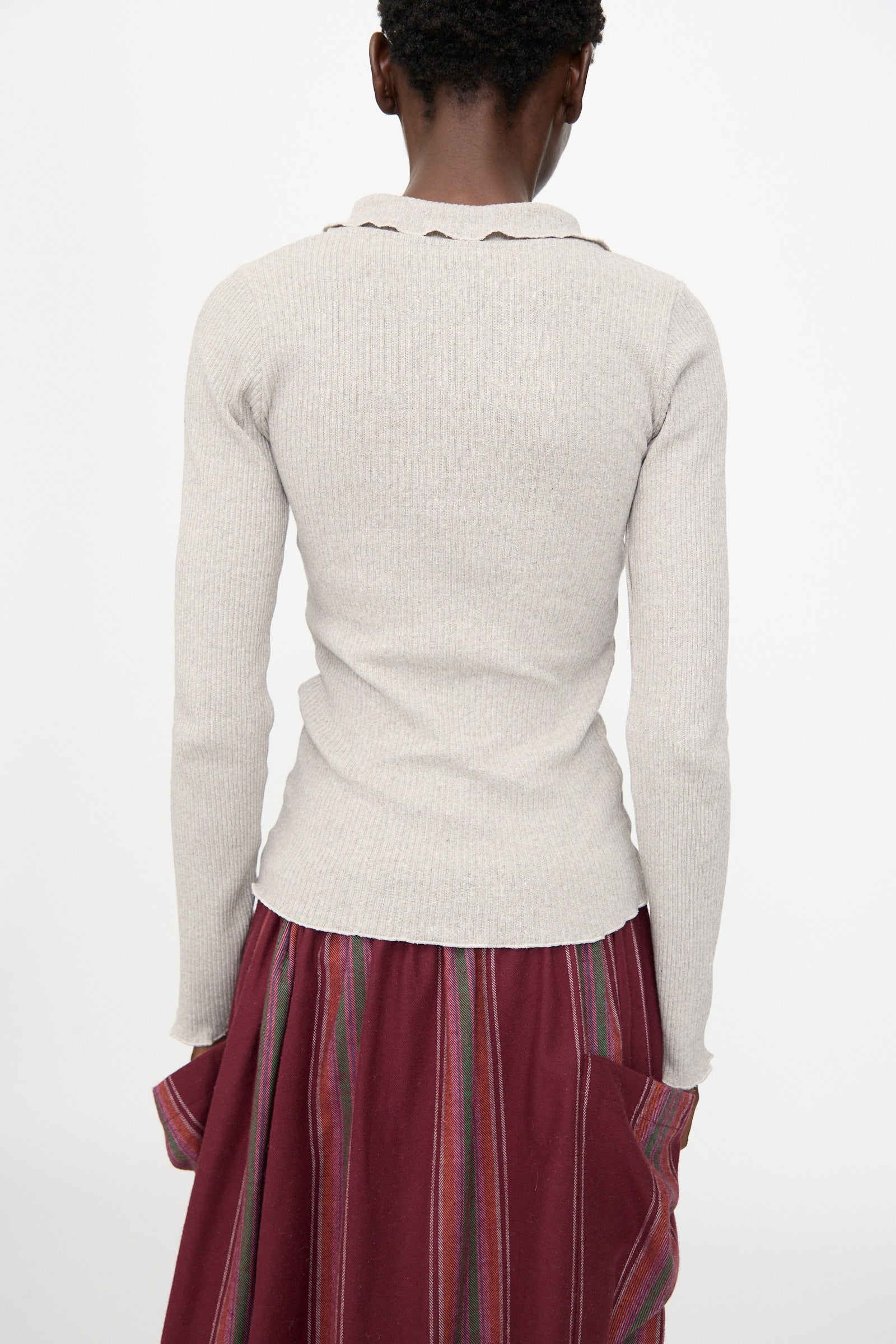 Person stands with their back to the camera, wearing the Baserange Dual Long Sleeve Tee in Grey Melange, a slim fit top made from recycled cotton, paired with a maroon skirt featuring multicolored vertical stripes.