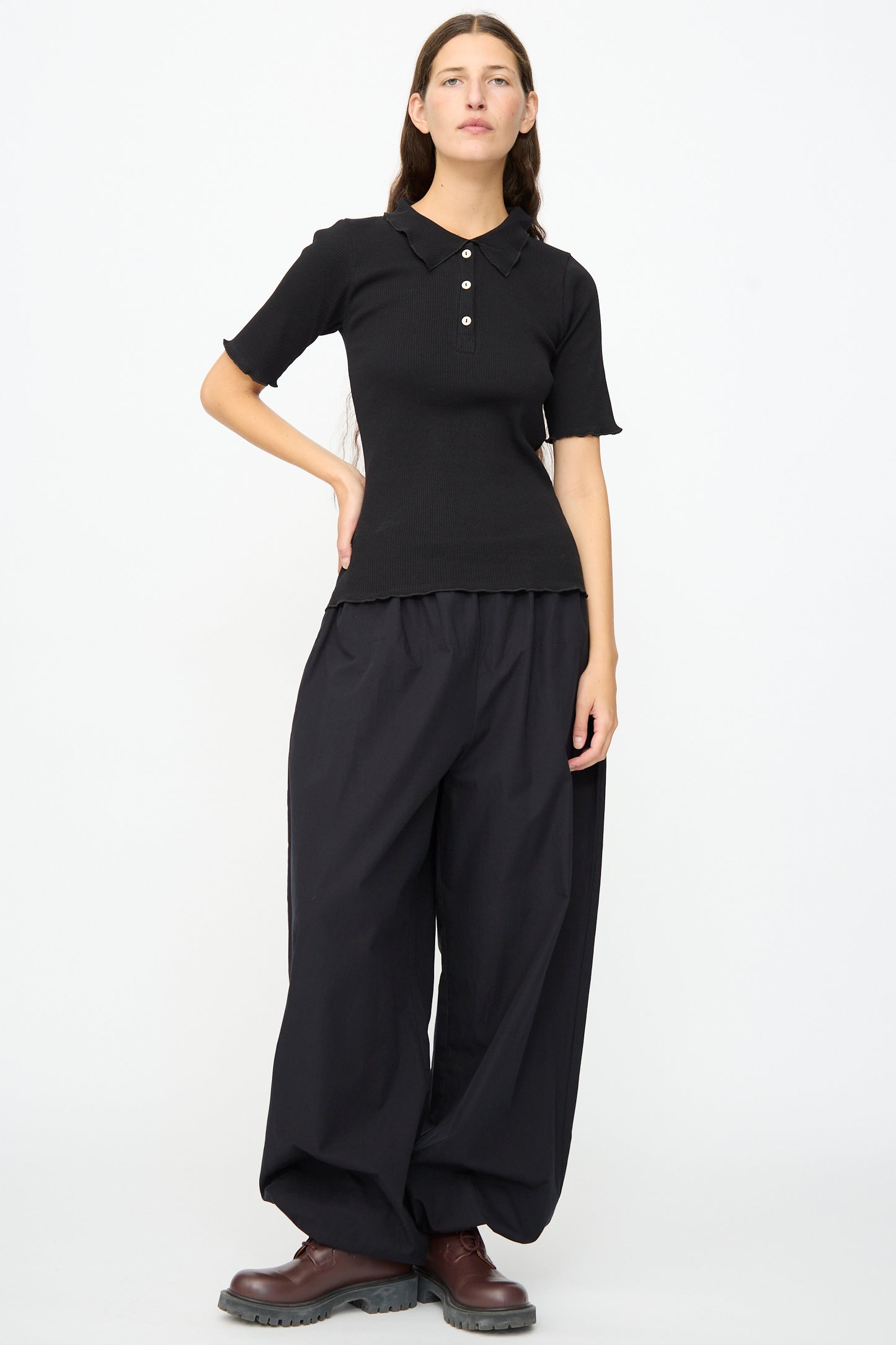 A person stands against a plain background wearing the Baserange Dual Polo in Black—a slim fit, short-sleeved shirt—paired with wide black pants and dark brown shoes, with one hand on their hip.