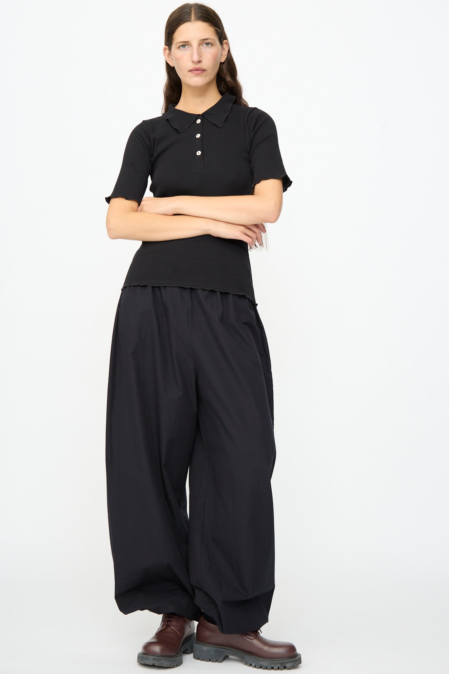 A person stands against a white background, wearing the Baserange Dual Polo in Black with ribbed short sleeves, paired with loose black pants and dark brown boots. Their arms are crossed and they have a neutral expression.