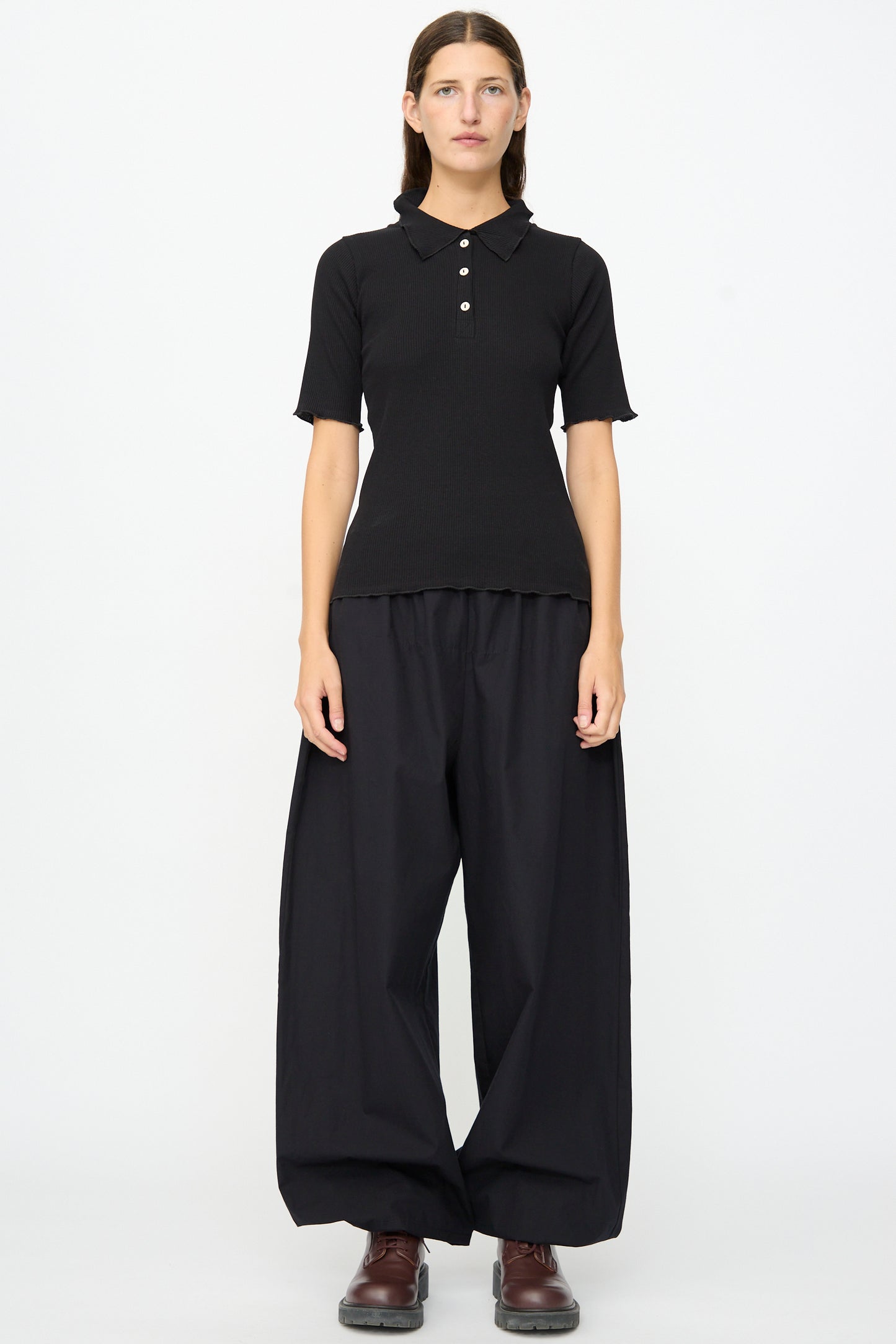 A person stands against a plain background wearing the Baserange Dual Polo in Black—a slim fit shirt with ribbed short sleeves—paired with loose black wide-leg pants and brown shoes.