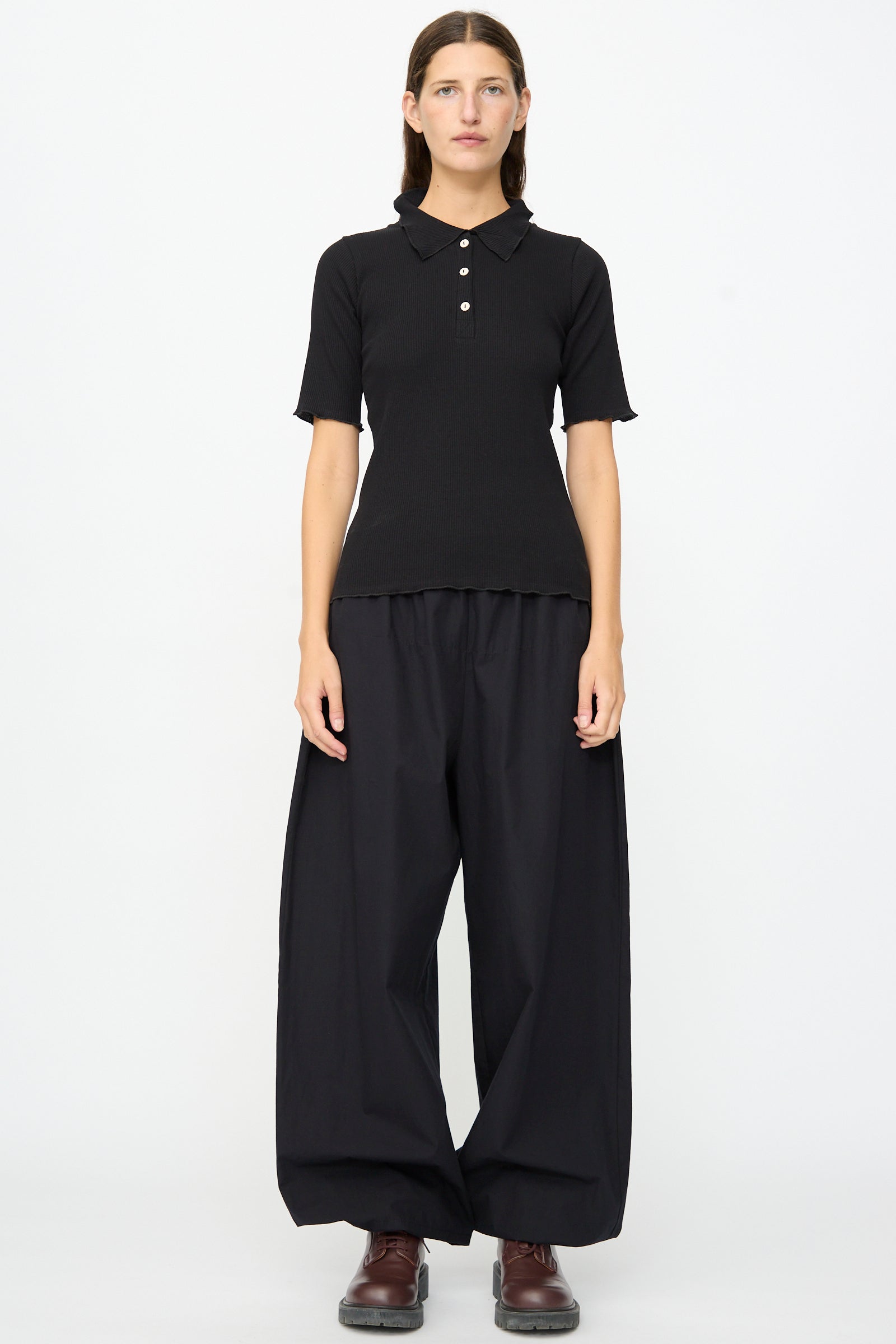 A person stands against a plain background wearing the Baserange Dual Polo in Black—a slim fit shirt with ribbed short sleeves—paired with loose black wide-leg pants and brown shoes.