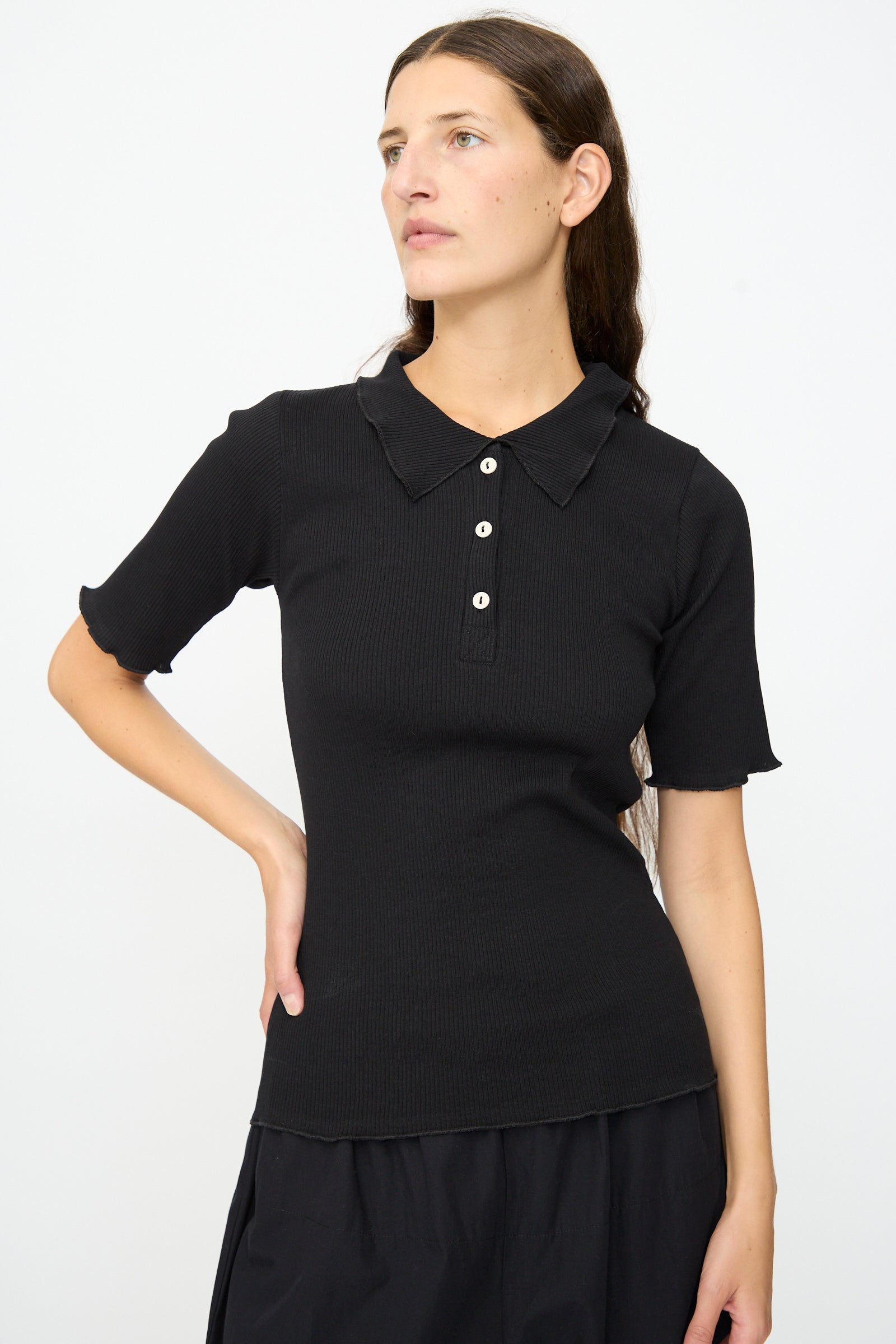 A woman with long brown hair is wearing the Baserange Dual Polo in Black—a slim fit, short-sleeved shirt—paired with a black skirt, standing against a plain white background.