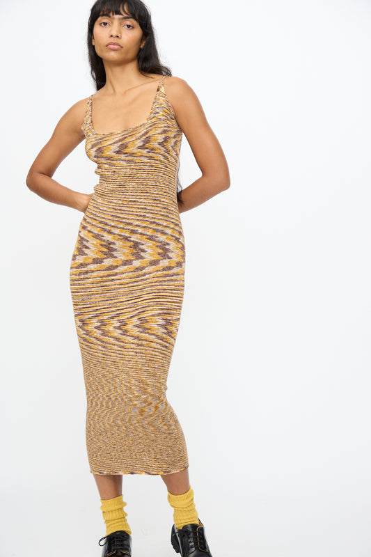 A woman stands against a plain white background wearing the Baserange Flute Dress in Brown Melange, paired with yellow socks and black shoes.