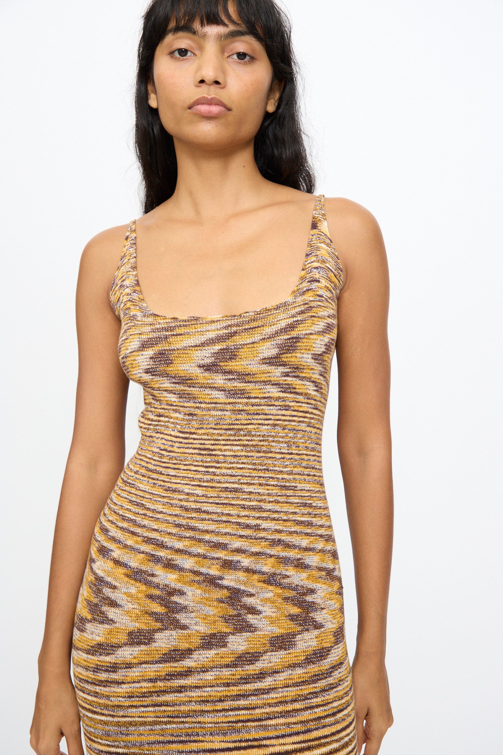 A woman with long dark hair wears the Baserange Flute Dress in Brown Melange, a slim-fit knit midi dress with a scooped neckline, standing against a plain white background.