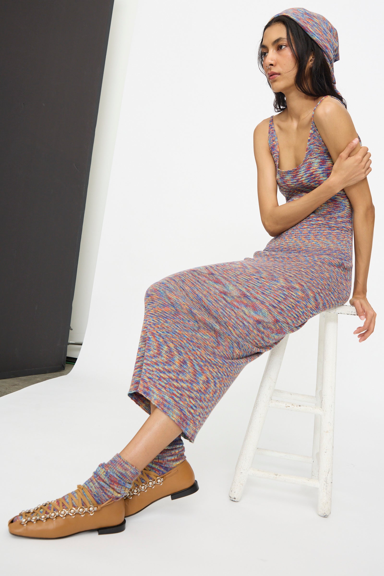 A woman in the Baserange Flute Dress in Multicolored, with a matching headscarf and socks, and tan chain-detail shoes, sits on a white stool against a plain background.