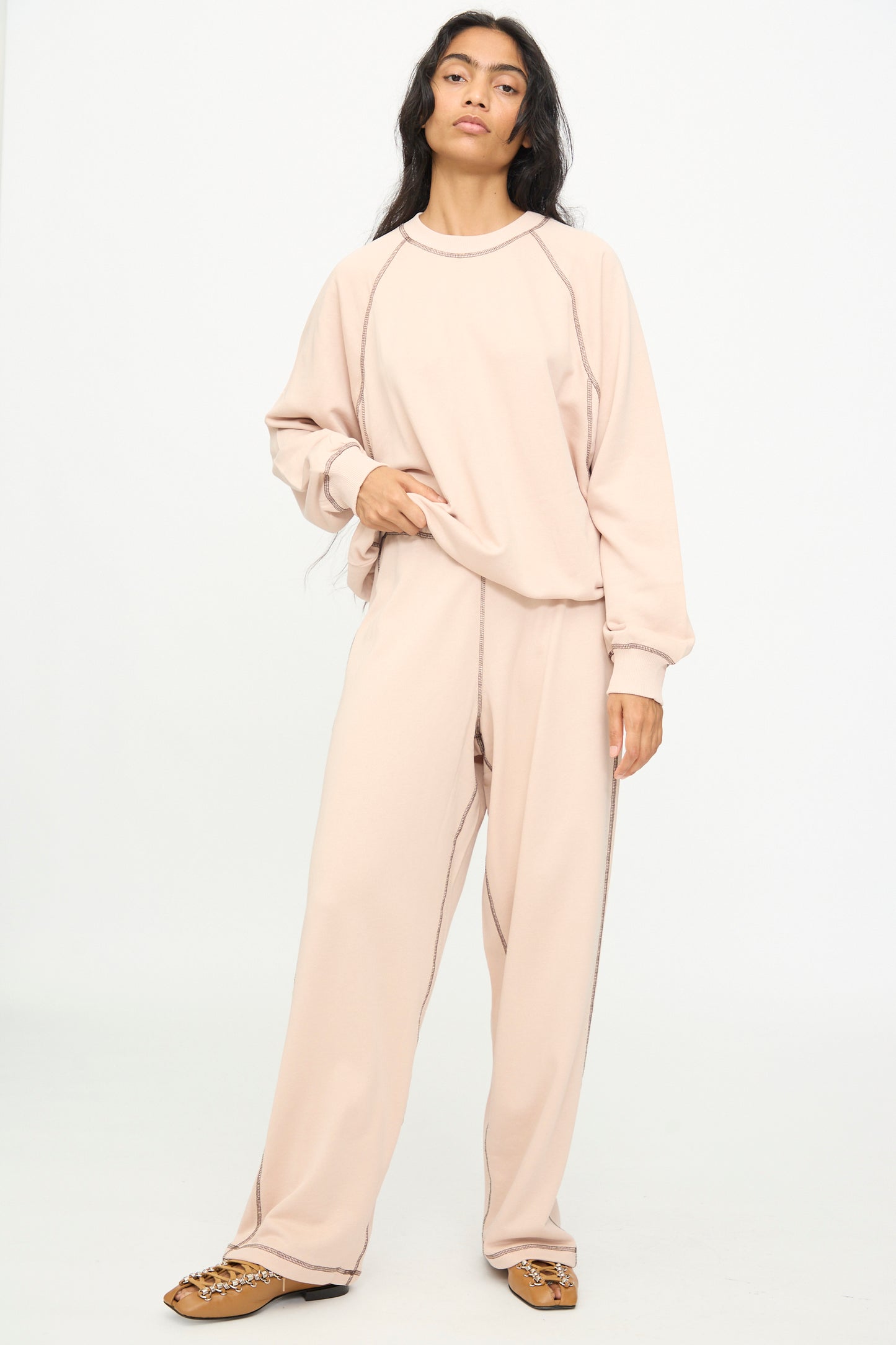 A woman stands against a white background wearing Baserange’s Fold Sweatpants in Haptic—a loose beige cotton blend style with a relaxed fit and elastic waistband—paired with a matching sweatshirt and tan loafers.