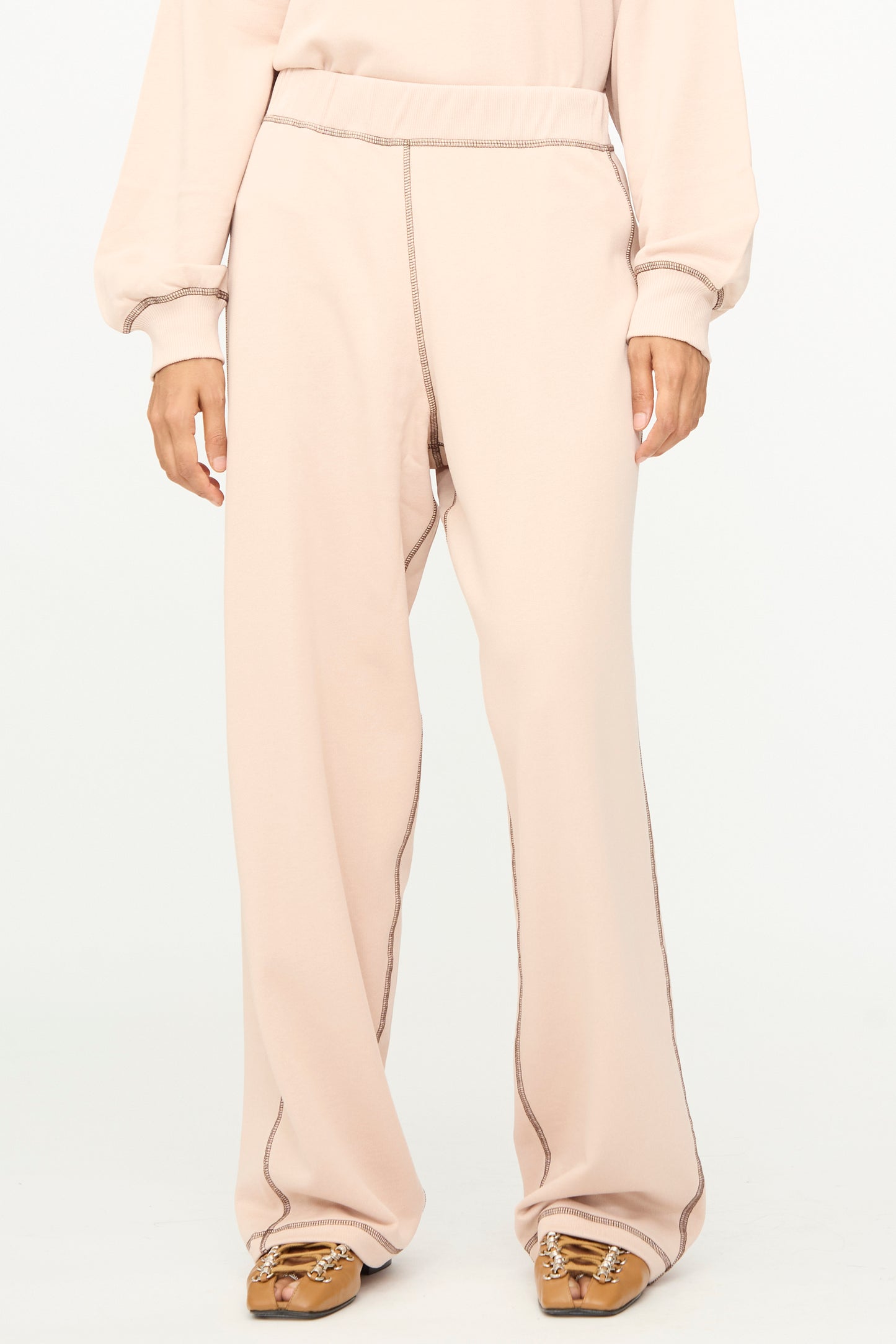 A person is shown from the shoulders down wearing Baserange Fold Sweatpants in Haptic—a loose, light beige, relaxed-fit style featuring an elastic waistband and visible seams—paired with tan moccasin-style shoes against a plain white background.