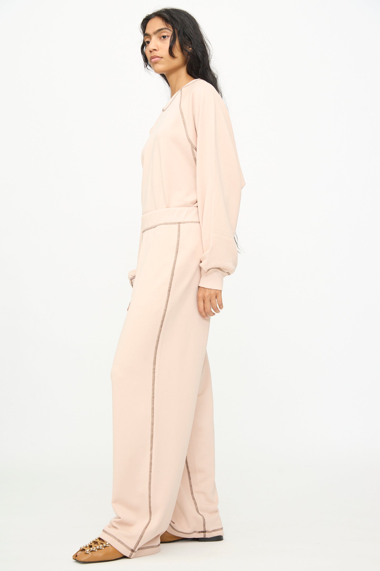 A woman stands sideways against a plain white background, wearing Baserange's Fold Sweatpants in Haptic—a relaxed style featuring an elastic waistband—paired with a light beige long-sleeve top and brown sandals.