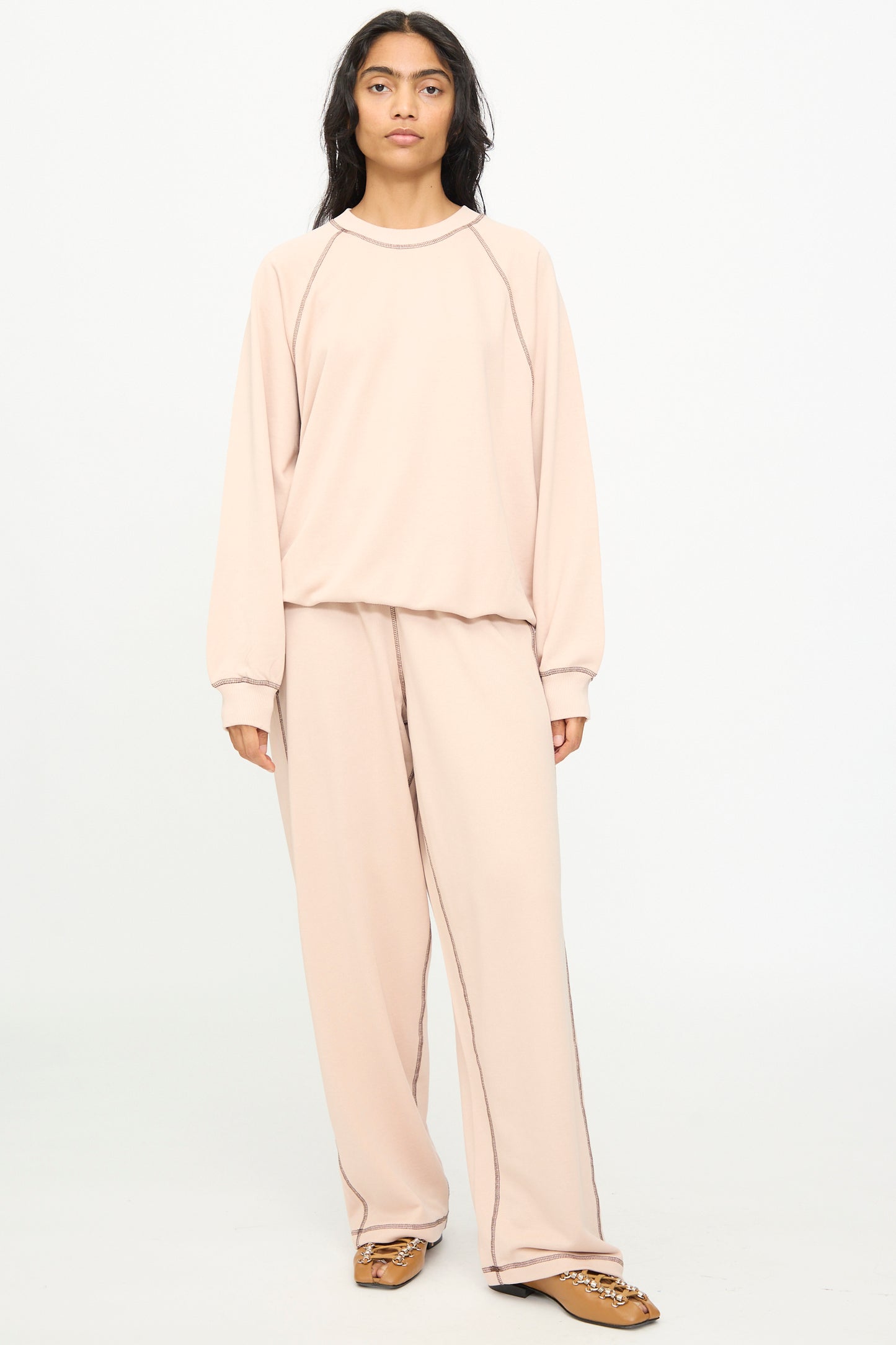 A person stands facing forward against a plain white background, wearing the Baserange Fold Sweatshirt in Haptic—a loose-fitting, light beige crewneck sweatshirt with raglan sleeves—paired with matching wide-leg pants and brown shoes.