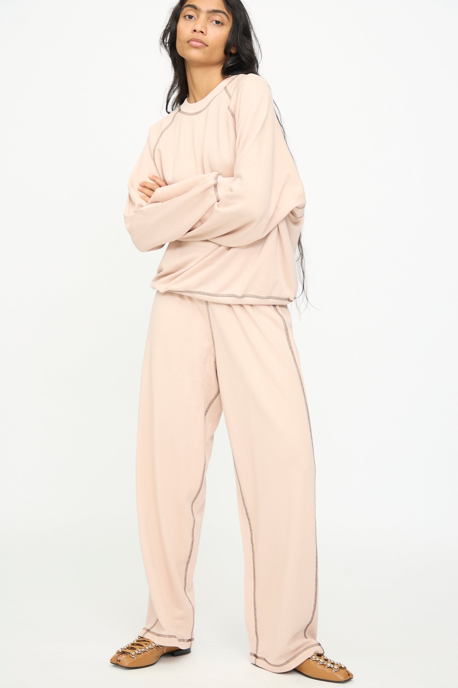 A person stands with arms crossed against a plain white background, wearing the Baserange Fold Sweatshirt in Haptic—a loose, light beige lyocell-cotton crewneck with raglan sleeves—matched with wide-leg pants and tan slip-on shoes.