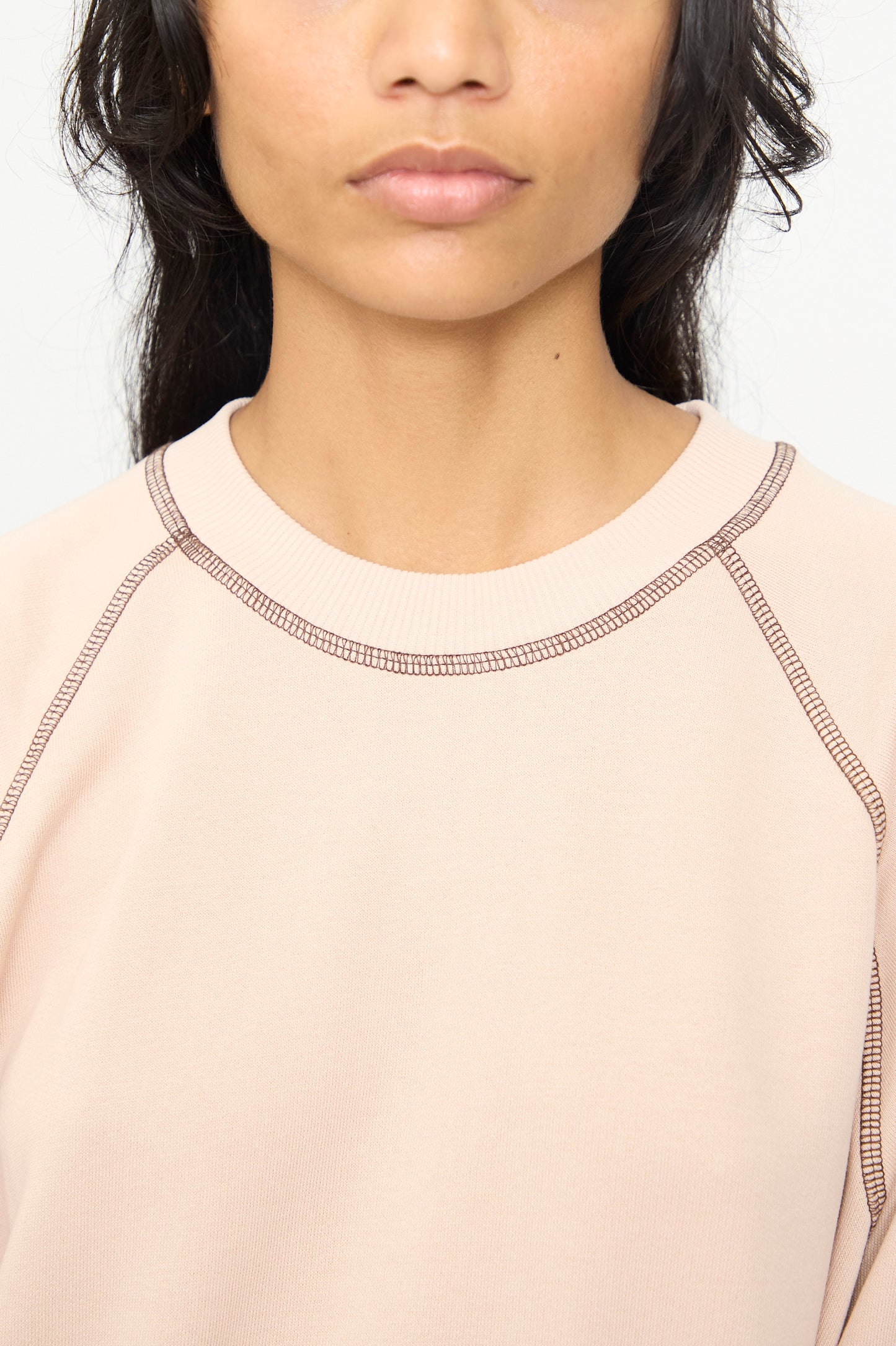 A person wearing the Baserange Fold Sweatshirt in Haptic—a light pink crewneck style with visible dark stitching and raglan sleeves—shown from the shoulders up with their face partially visible.