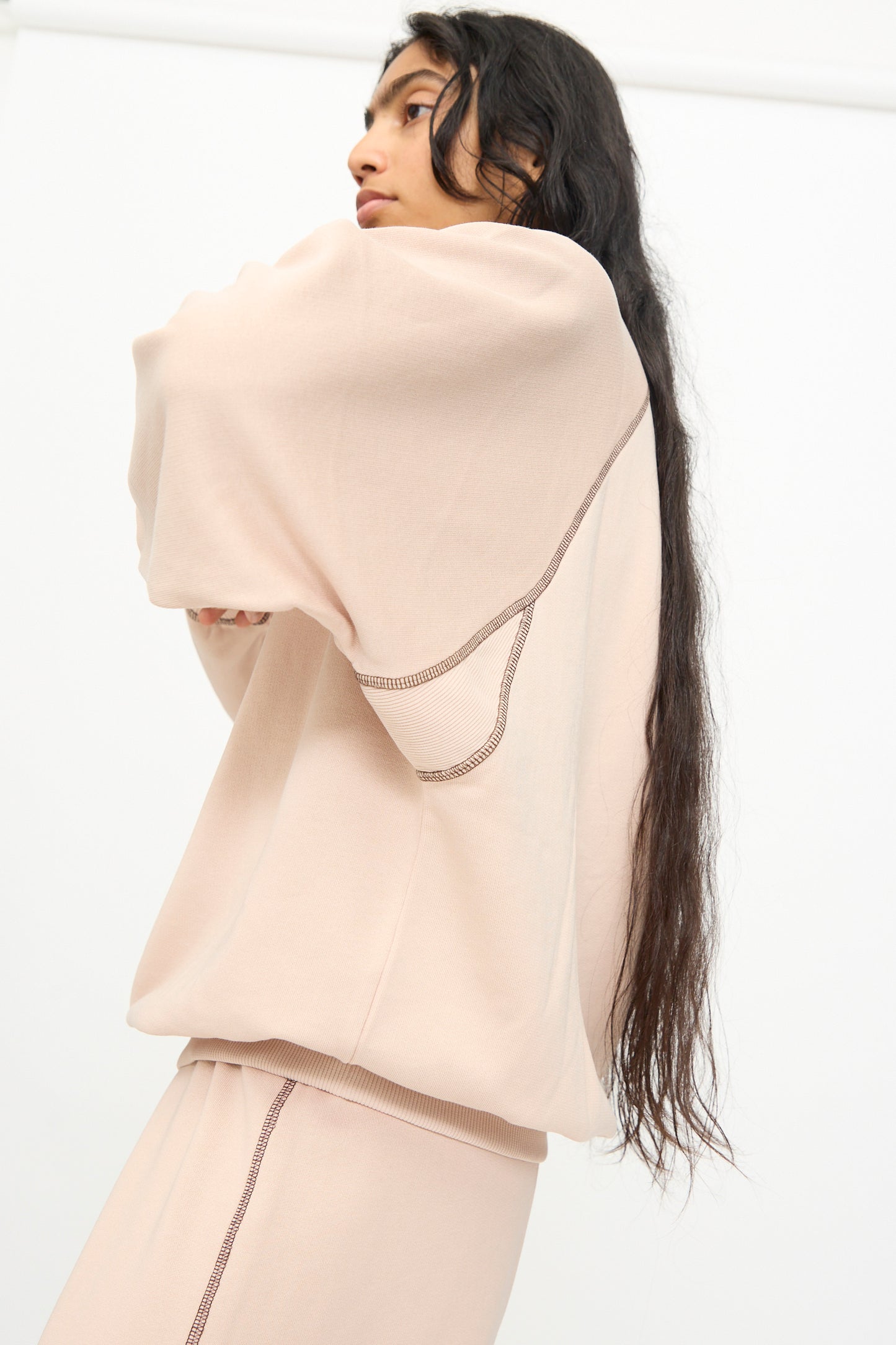 A person with long dark hair wears the Baserange Fold Sweatshirt in Haptic—a loose, light beige lyocell cotton sweatshirt featuring raglan sleeves and visible stitching—standing against a plain white background, facing slightly away from the camera.