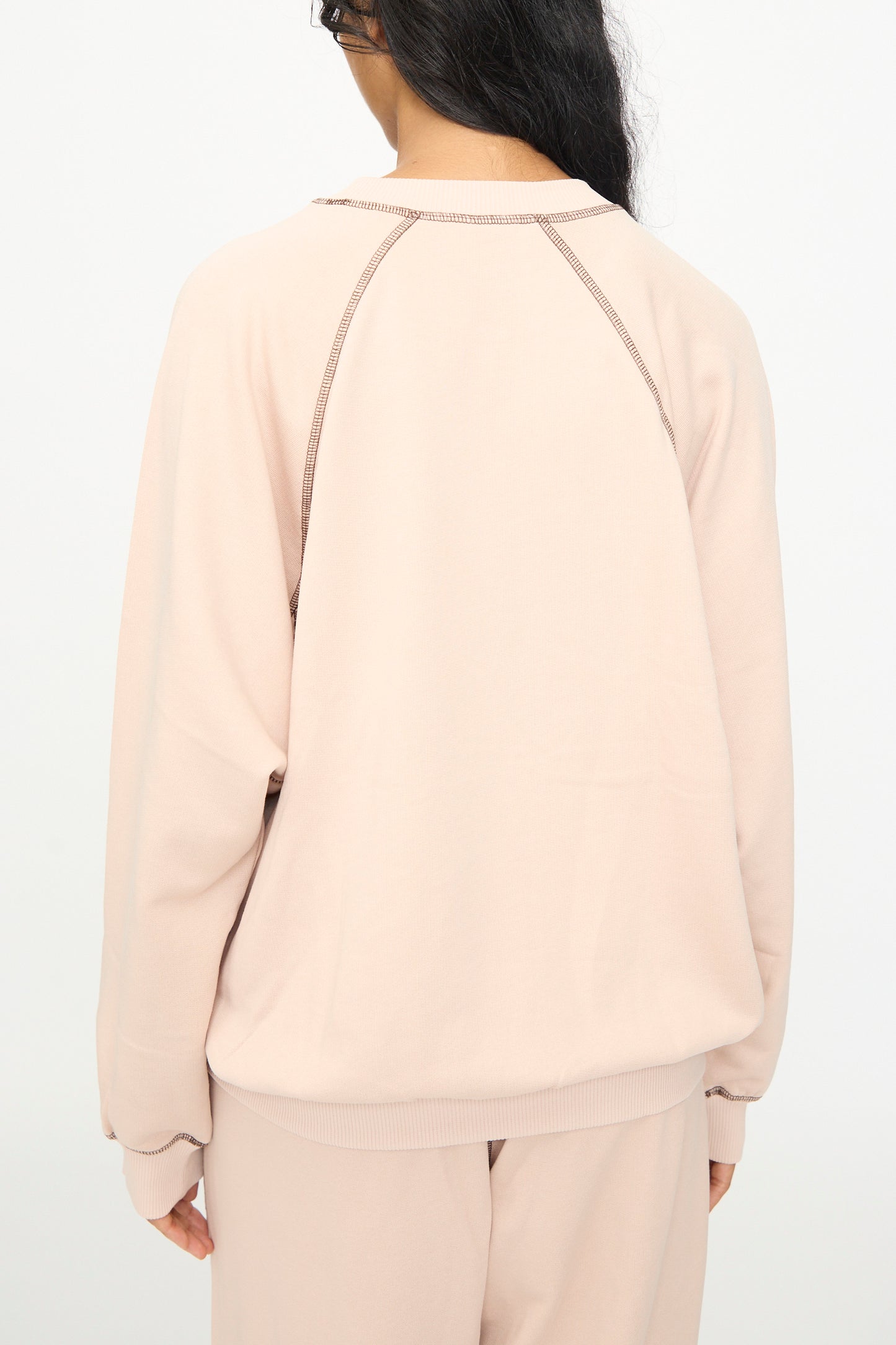 A person with long dark hair is seen from the back, wearing the Baserange Fold Sweatshirt in Haptic—a light beige crewneck style with raglan sleeves—paired with matching pants, standing against a plain white background.