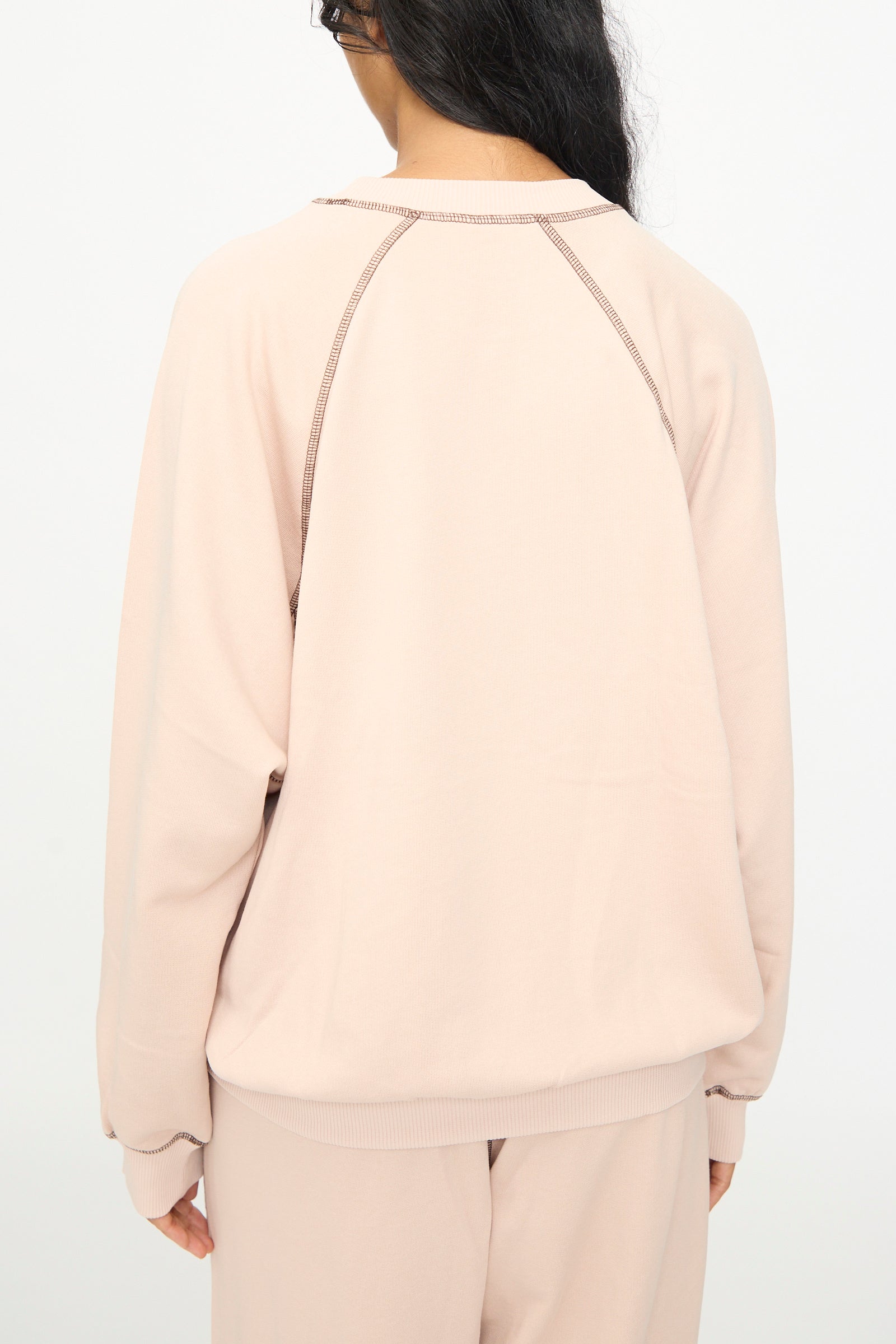 A person with long dark hair is seen from the back, wearing the Baserange Fold Sweatshirt in Haptic—a light beige crewneck style with raglan sleeves—paired with matching pants, standing against a plain white background.