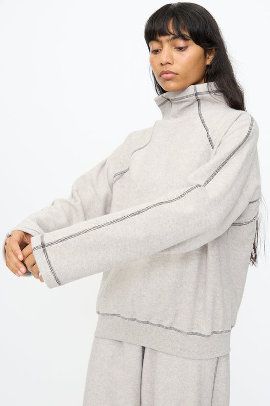 A person wears the Baserange Fold Turtleneck in Grey Melange, an oversized recycled cotton sweater with exposed seams, paired with light gray pants, standing against a plain white background.