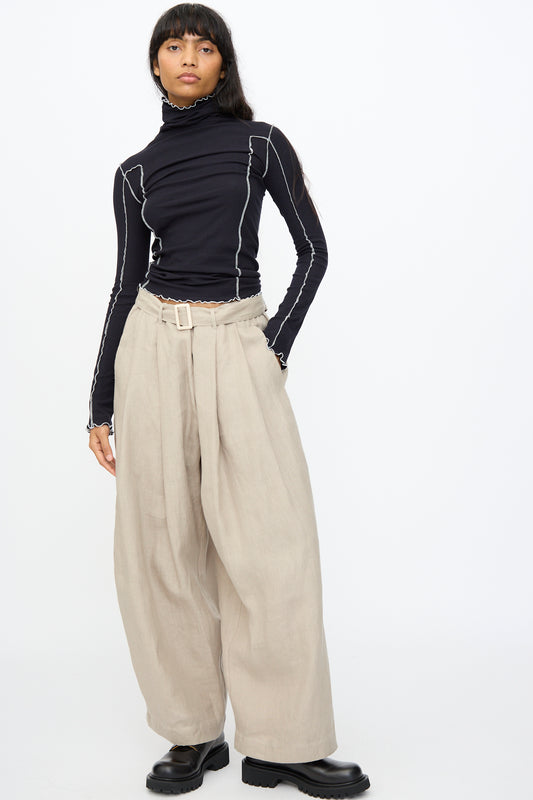 A person wearing a black long-sleeve turtleneck with white seams, Baserange Frame Pants in Concrete, and black shoes stands against a plain white background.