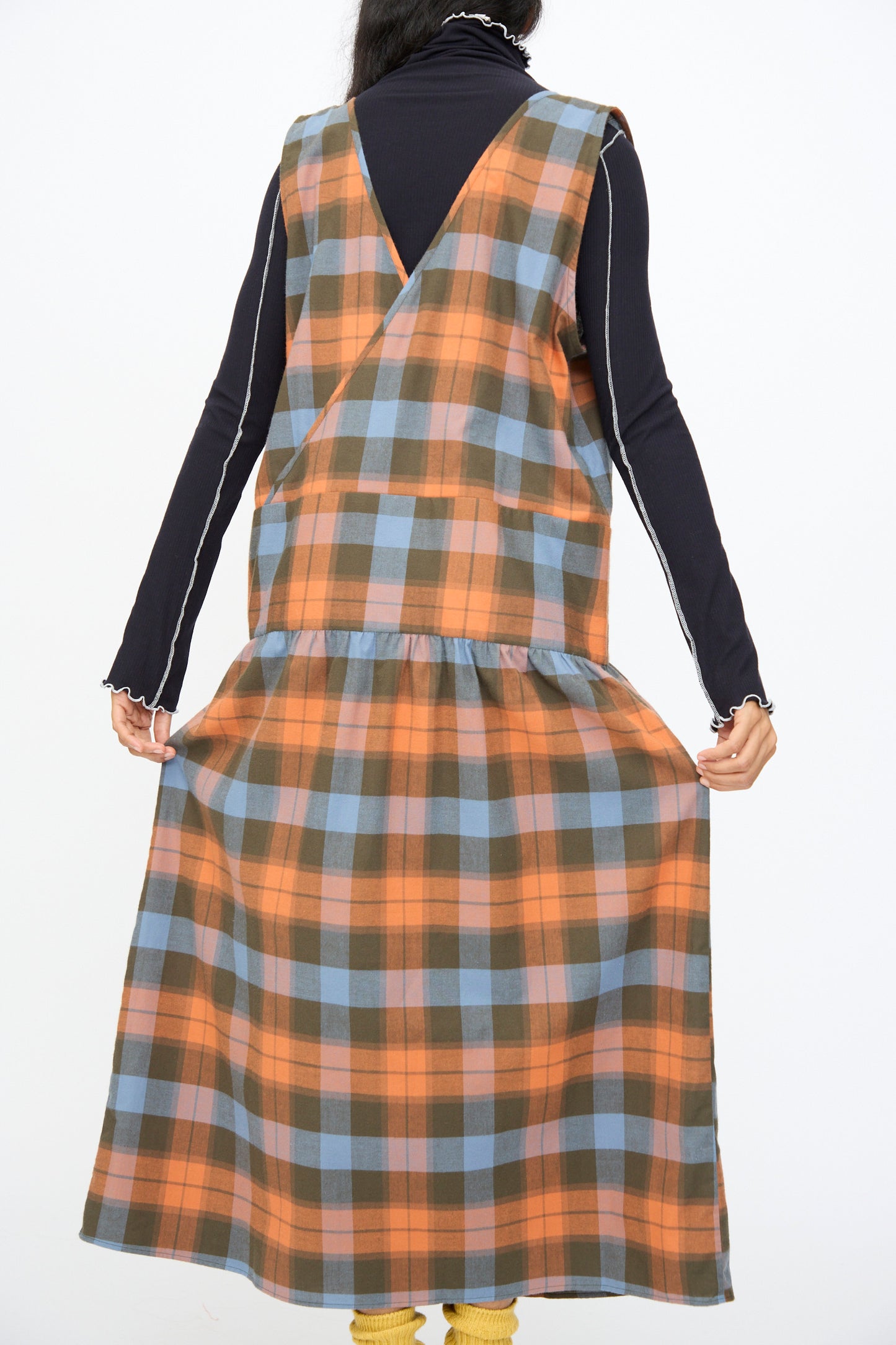 A person is shown from the back holding the sides of the Baserange Garlin Dress in Frame Plaid—an orange, blue, and brown organic cotton sleeveless dress—worn over a long-sleeve black top.