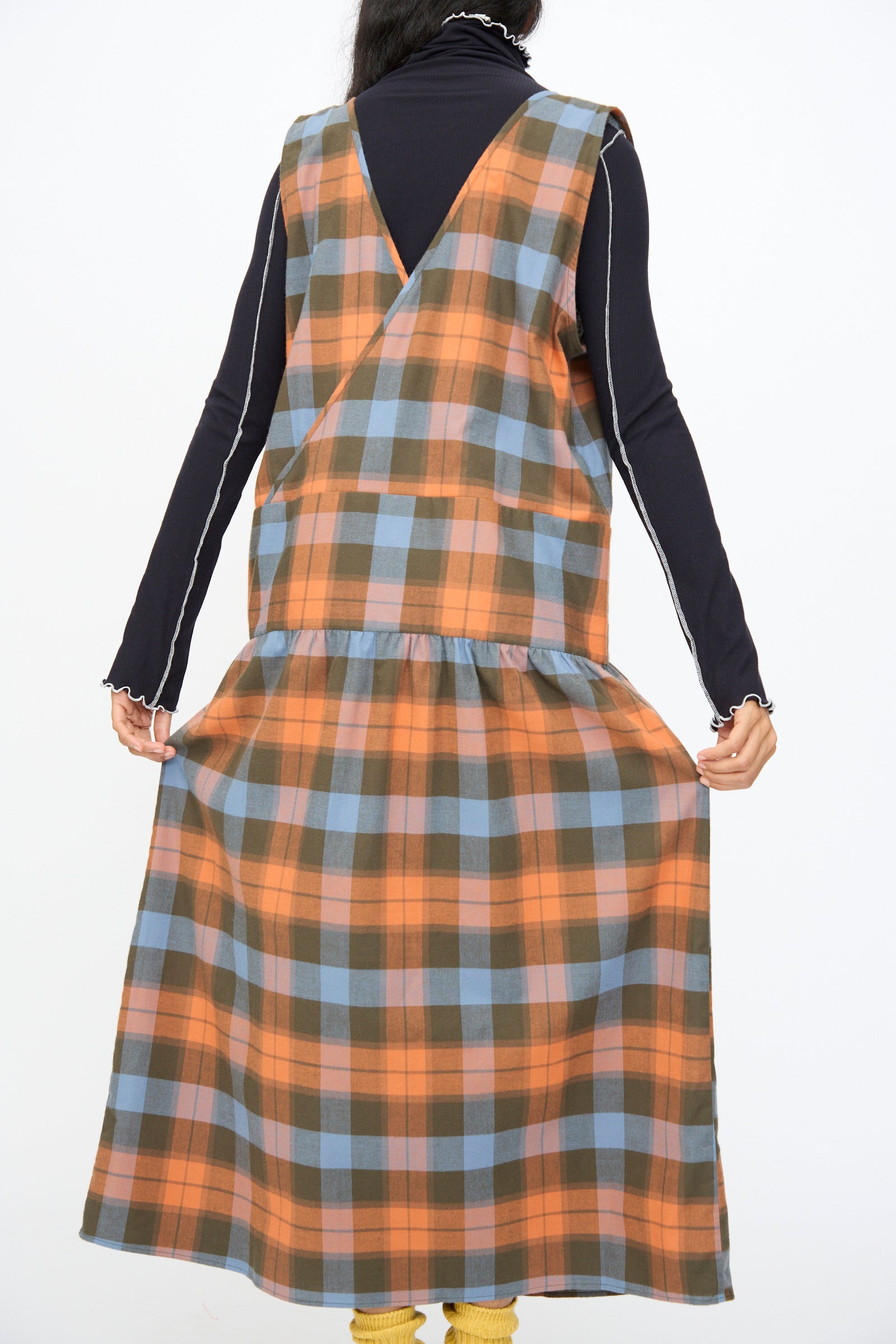 A person is shown from the back holding the sides of the Baserange Garlin Dress in Frame Plaid—an orange, blue, and brown organic cotton sleeveless dress—worn over a long-sleeve black top.