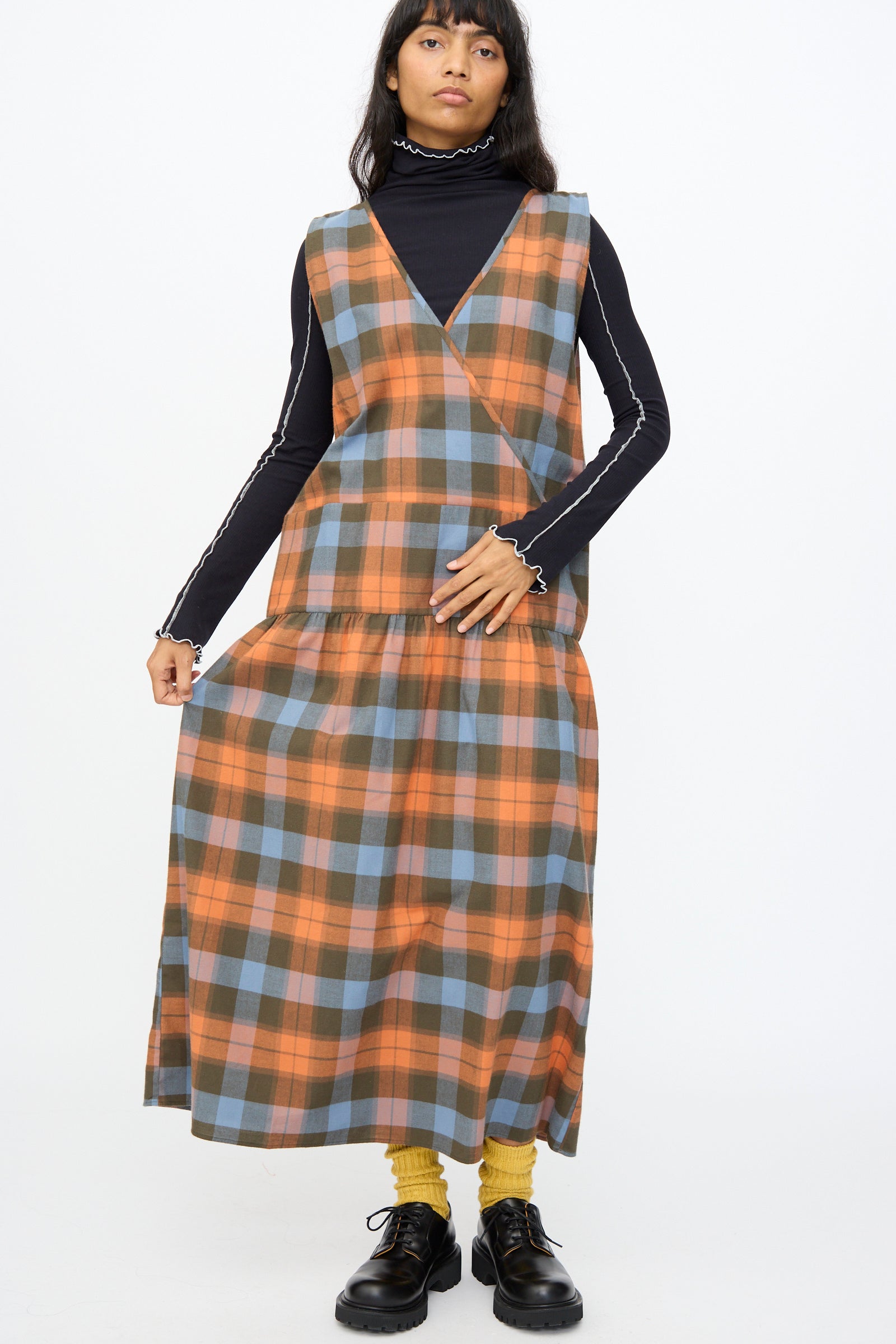 A person stands against a plain white background, wearing the Baserange Garlin Dress in Frame Plaid over a black turtleneck, paired with yellow socks and black shoes.