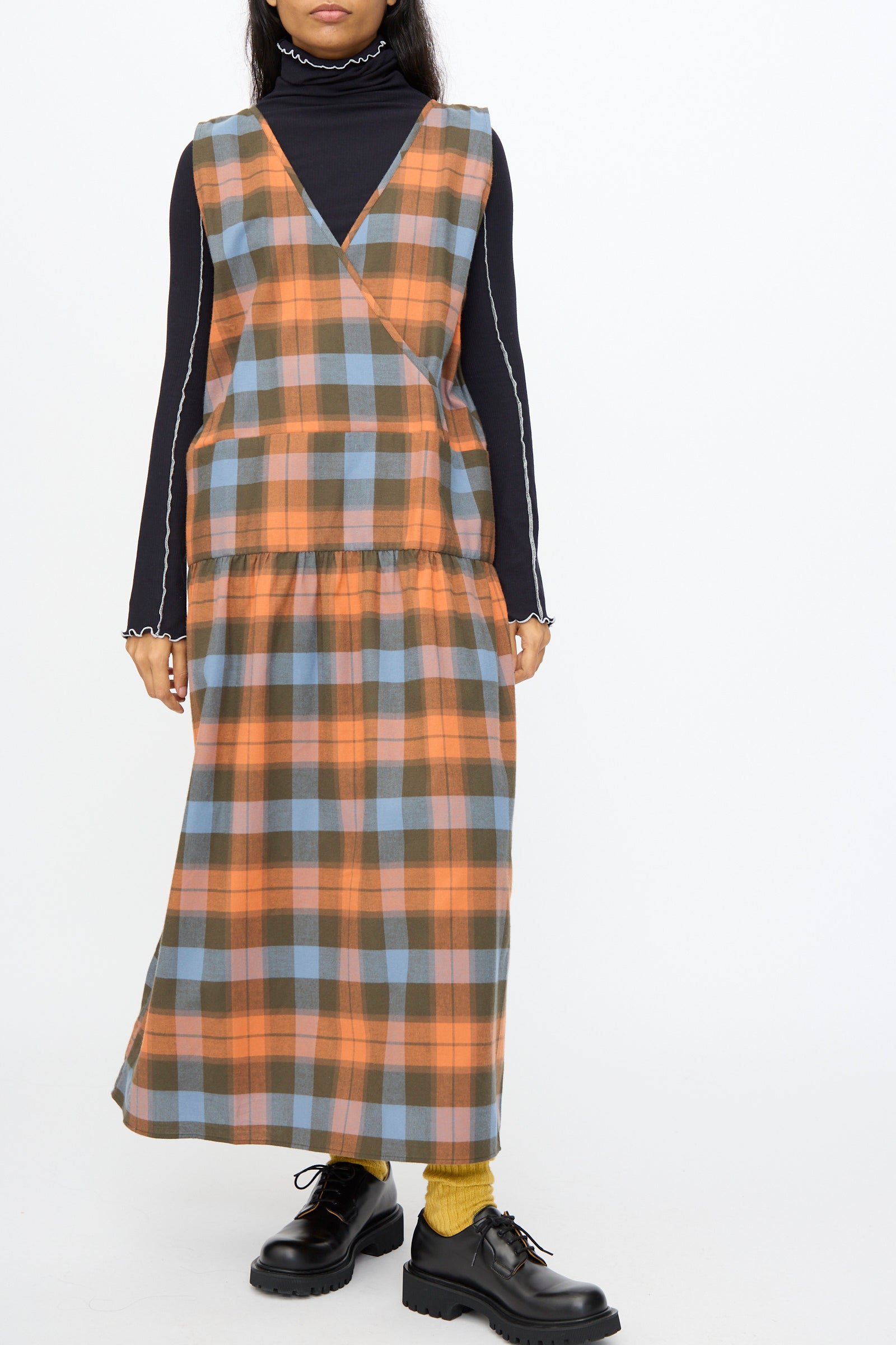 A person in the Baserange Garlin Dress in Frame Plaid—a sleeveless orange and blue plaid organic cotton dress with a gathered skirt—layered over a black turtleneck, yellow socks, and black chunky shoes, against a white background.
