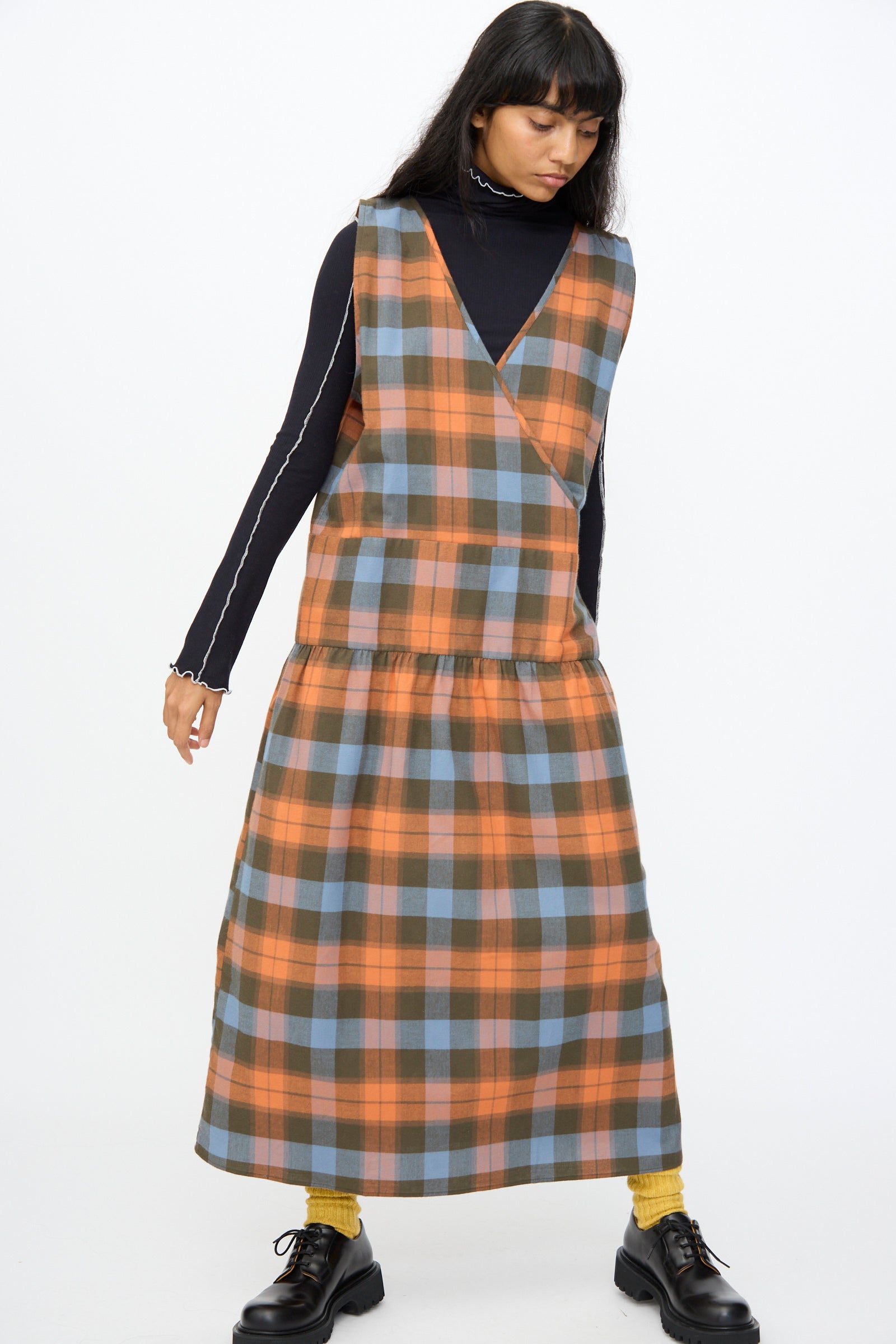 A woman wears the Baserange Garlin Dress in Frame Plaid—an orange and blue sleeveless style—over a black long-sleeve top with yellow socks and black shoes, standing against a plain white background. 