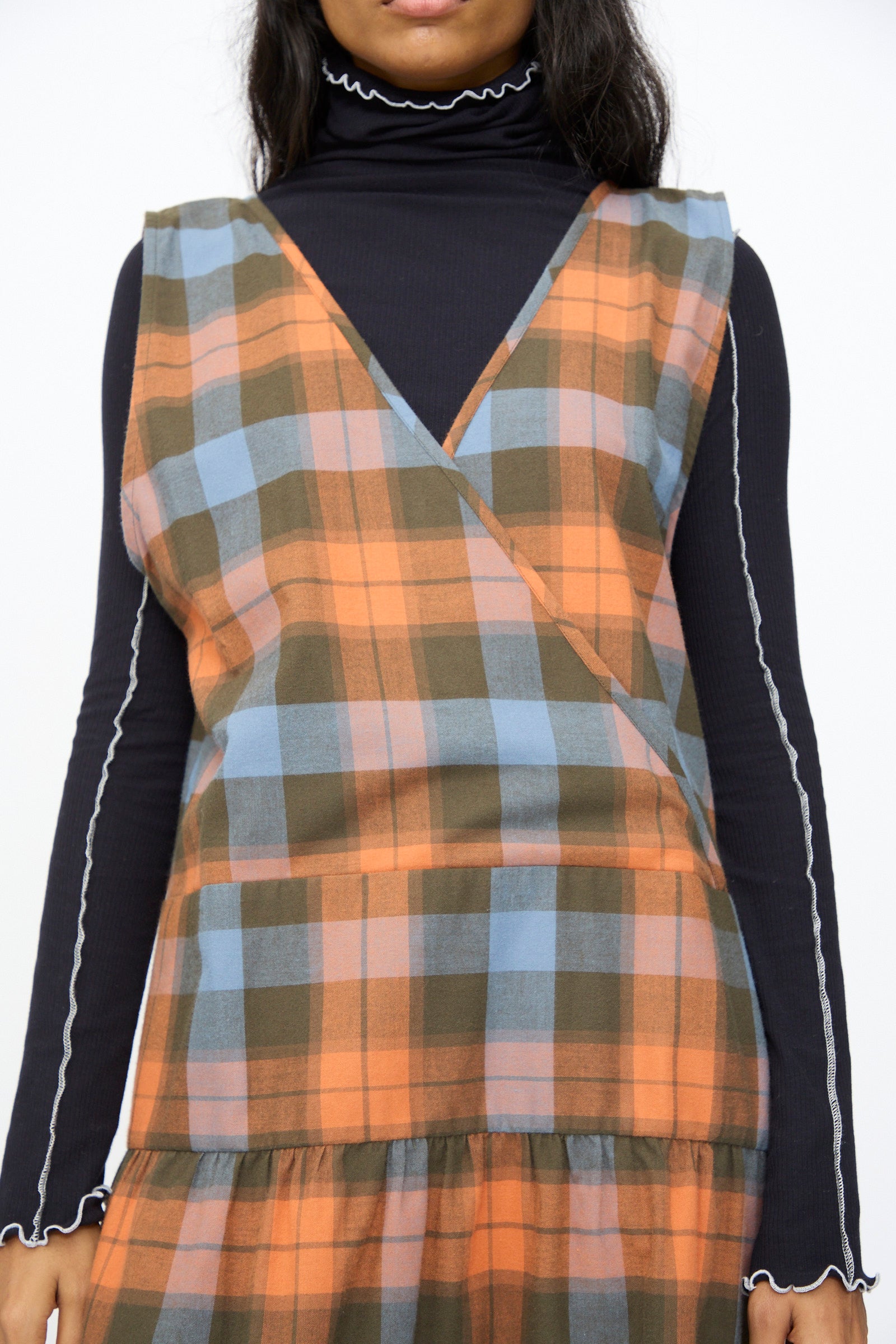 A person wears the Baserange Garlin Dress in Frame Plaid, an orange, blue, and brown plaid sleeveless dress made from soft organic cotton, styled over a black turtleneck with white trim for comfort and sustainability.